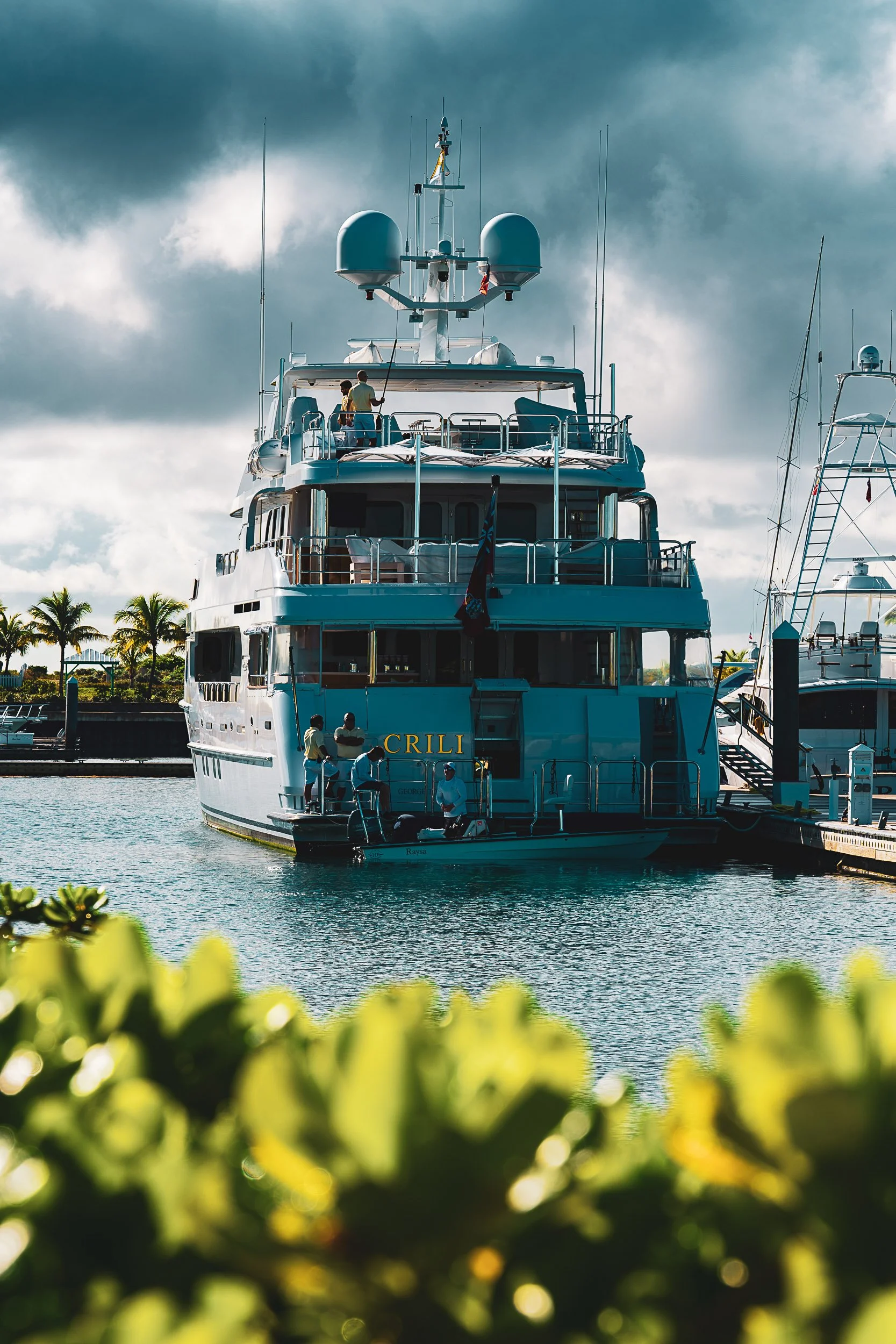 A large yacht docked at a marina with lush greenery and cloudy skies in the background.