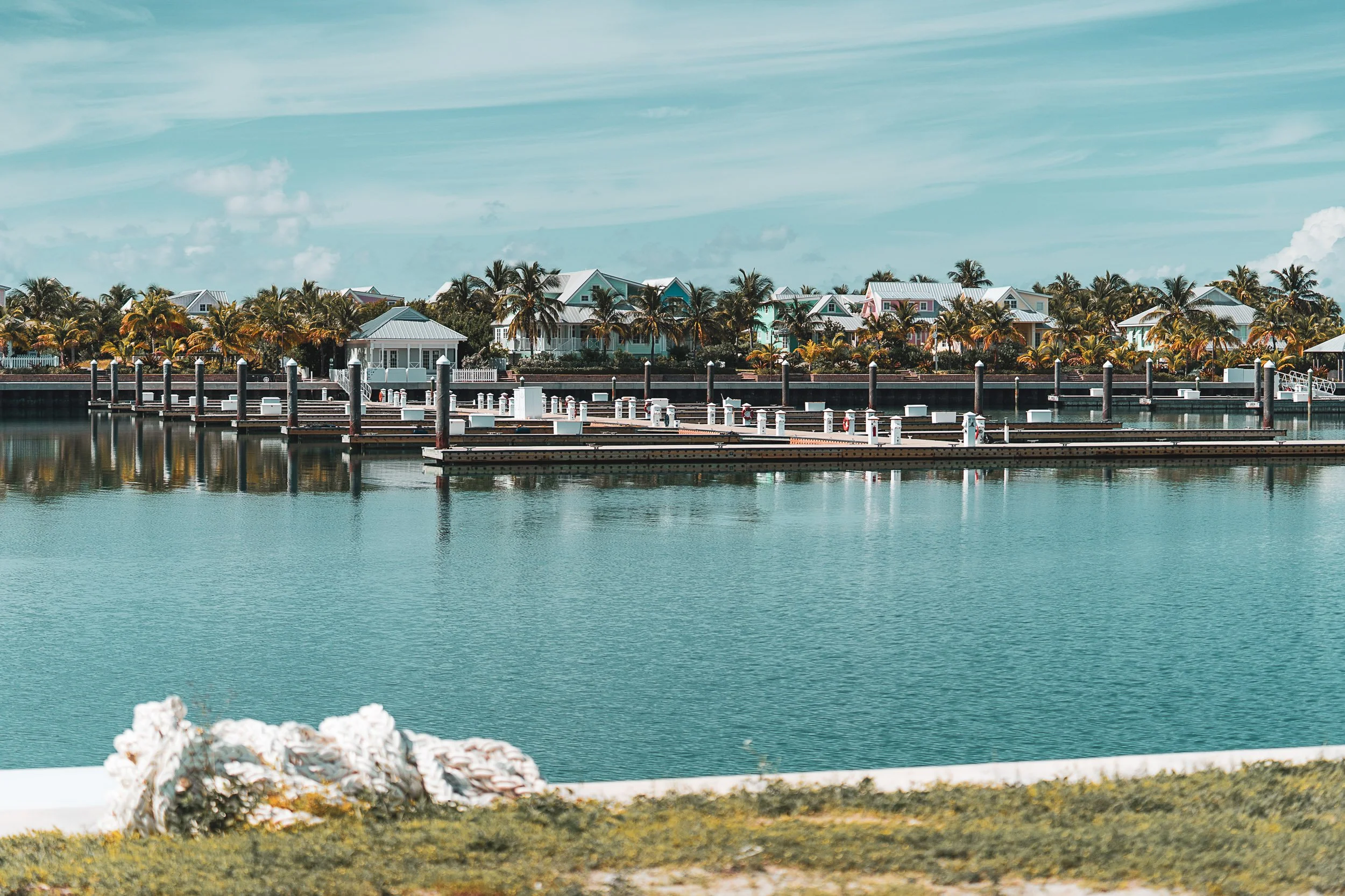 Scenic view of a waterfront marina with docks, surrounded by colorful houses and palm trees under a blue sky.