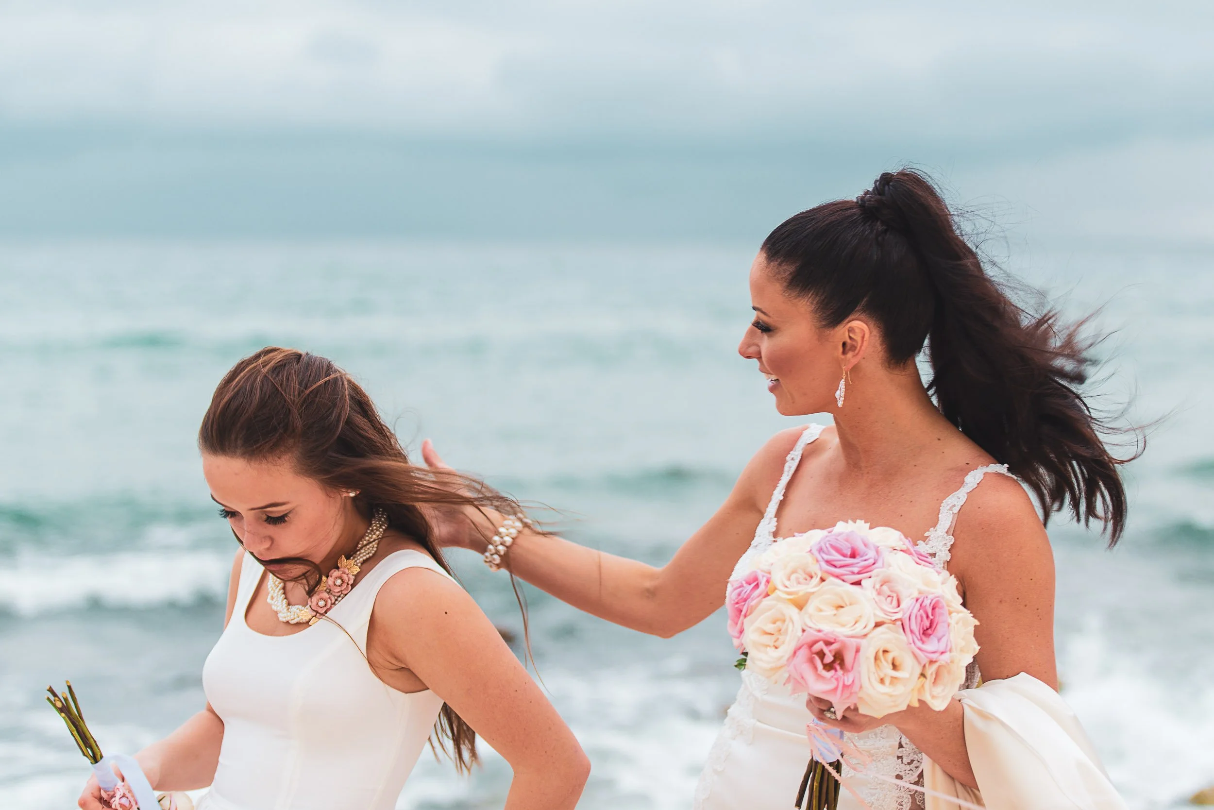 Two women in white dresses by the ocean, with one holding a bouquet of pink and white roses.