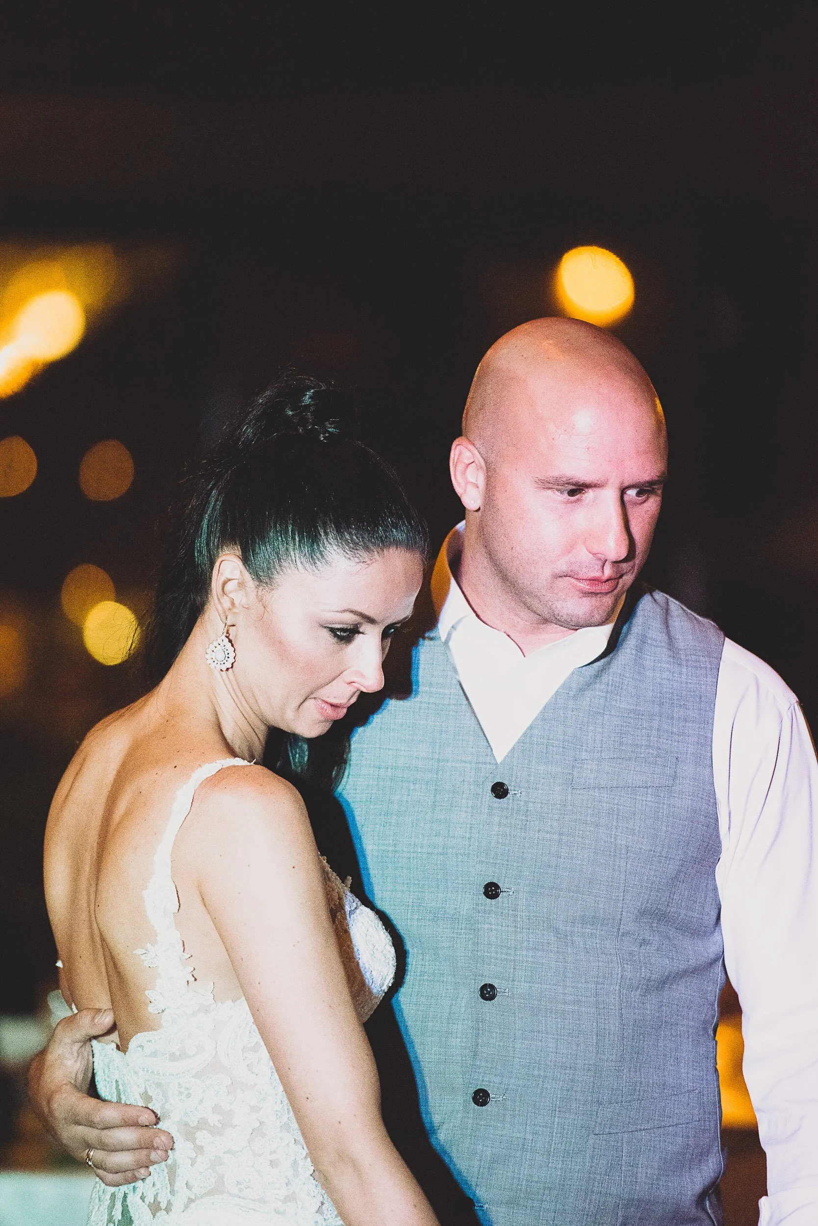 A couple embracing at a nighttime event, with the woman in a lace dress and the man wearing a gray vest.