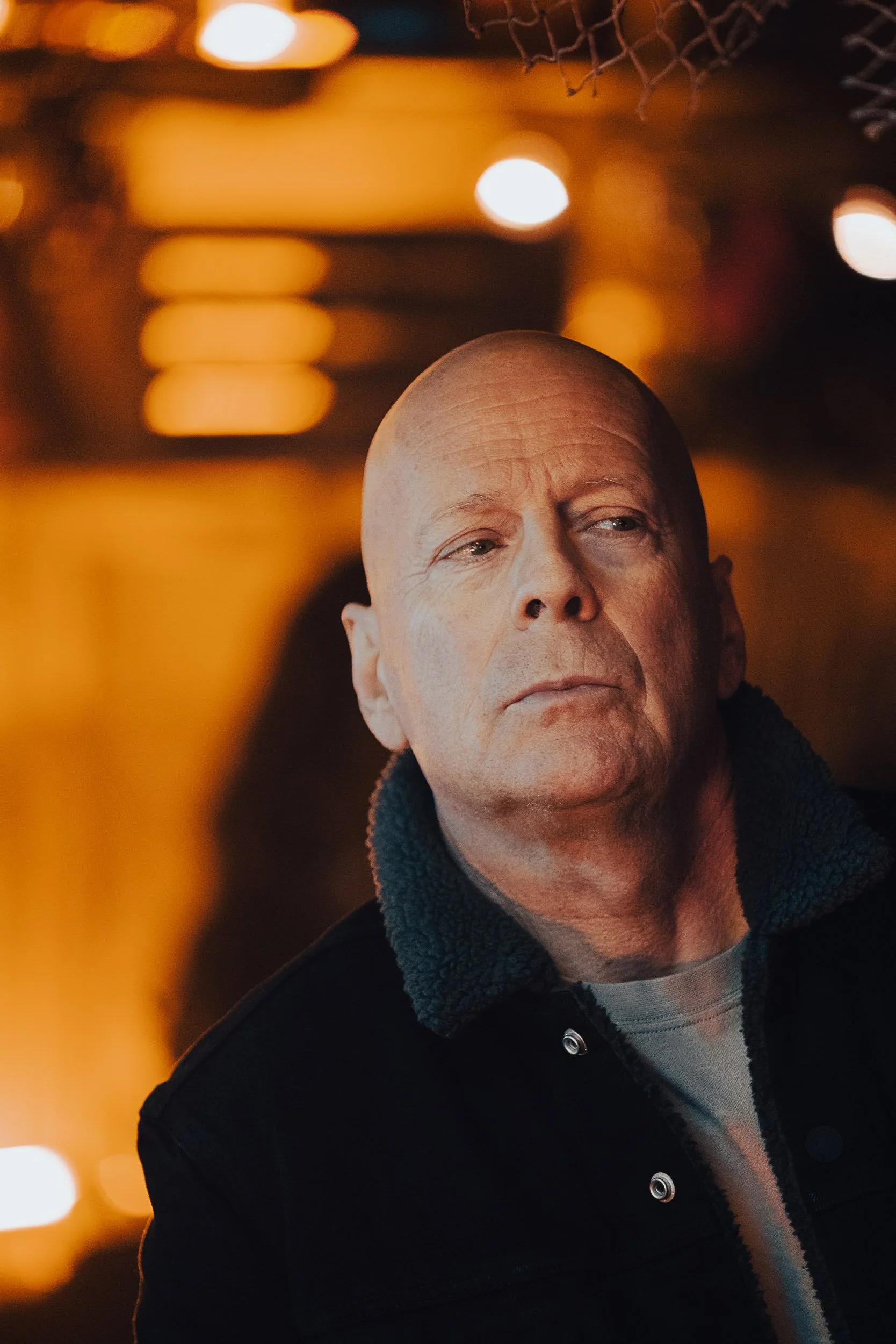 A man in a dark jacket with a fur collar, looking serious, stands in a dimly lit room with warm, blurred lights in the background.