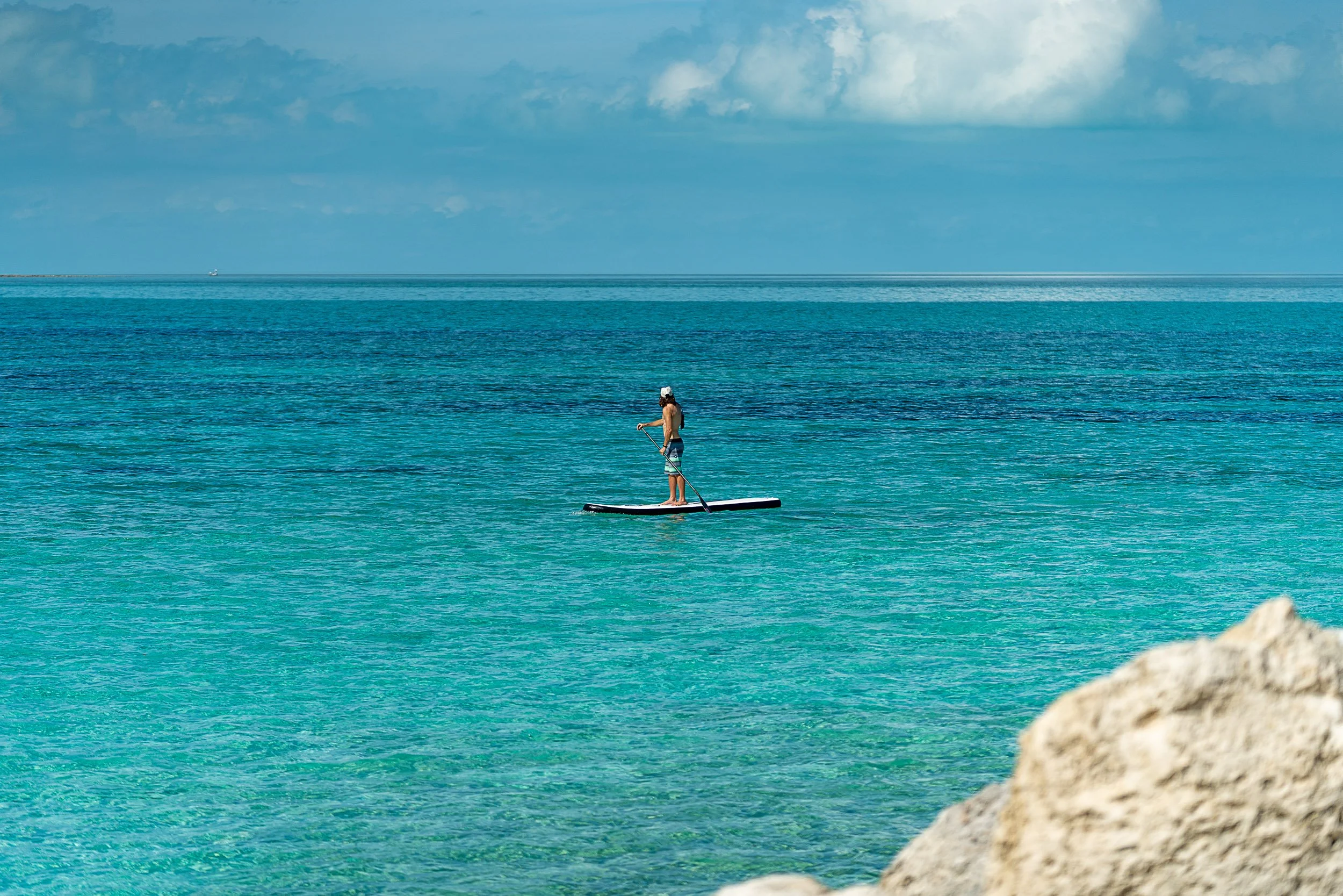 Person paddleboarding on calm, turquoise ocean water with blue sky and clouds in the background.