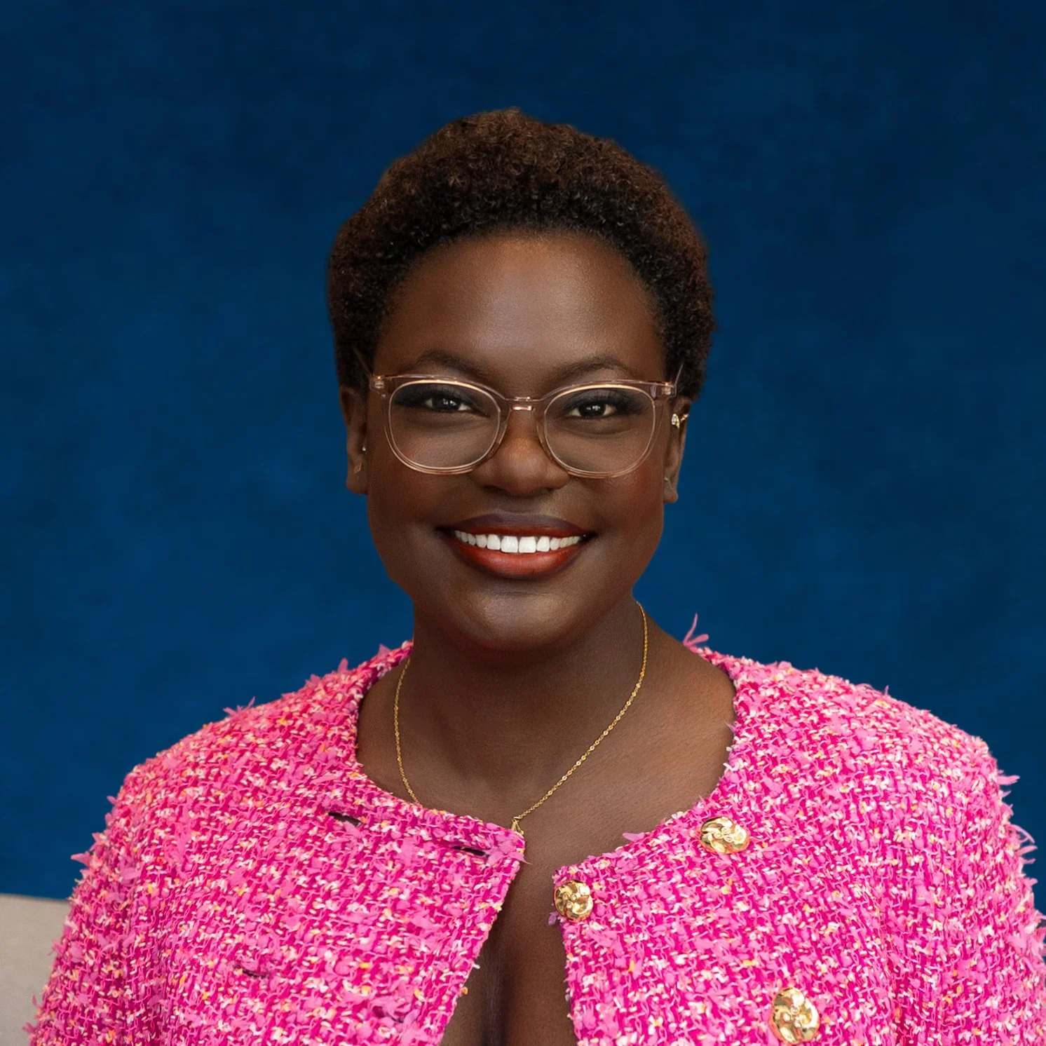 A woman with short curly hair, glasses, and a bright pink textured blazer with gold buttons, smiling against a blue background.