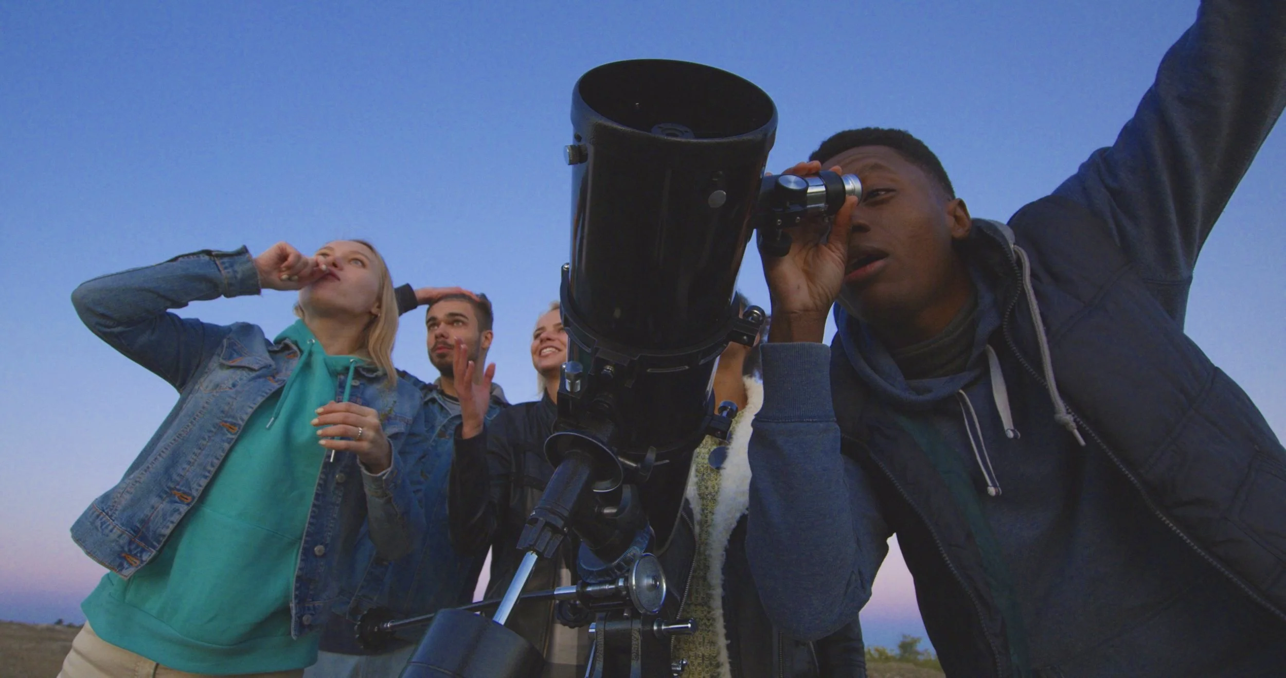 A group of young people outdoors during twilight, with one person looking through a telescope and others observing, talking, or making gestures.