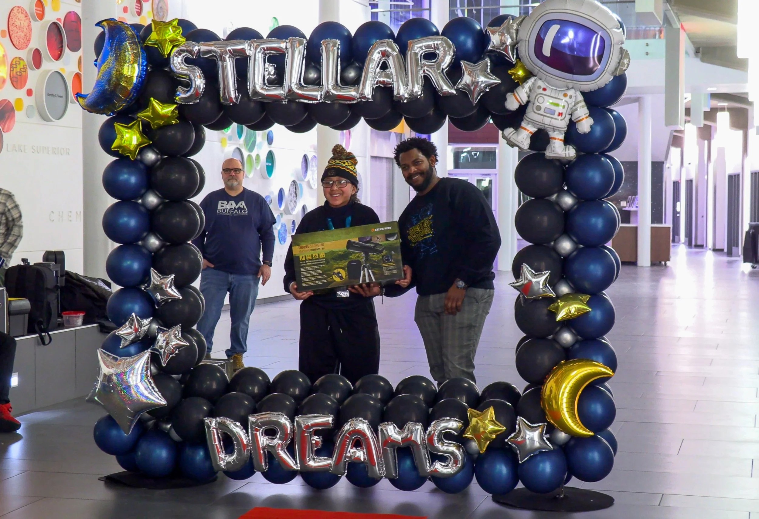 Three people standing inside a balloon frame with the words 'Stellar Dreams' spelled out in balloons. The person in the middle is holding a boxed toy, and the person on the right is smiling. In the background, a man in a blue sweatshirt stands. The frame is decorated with star-shaped and moon-shaped balloons.