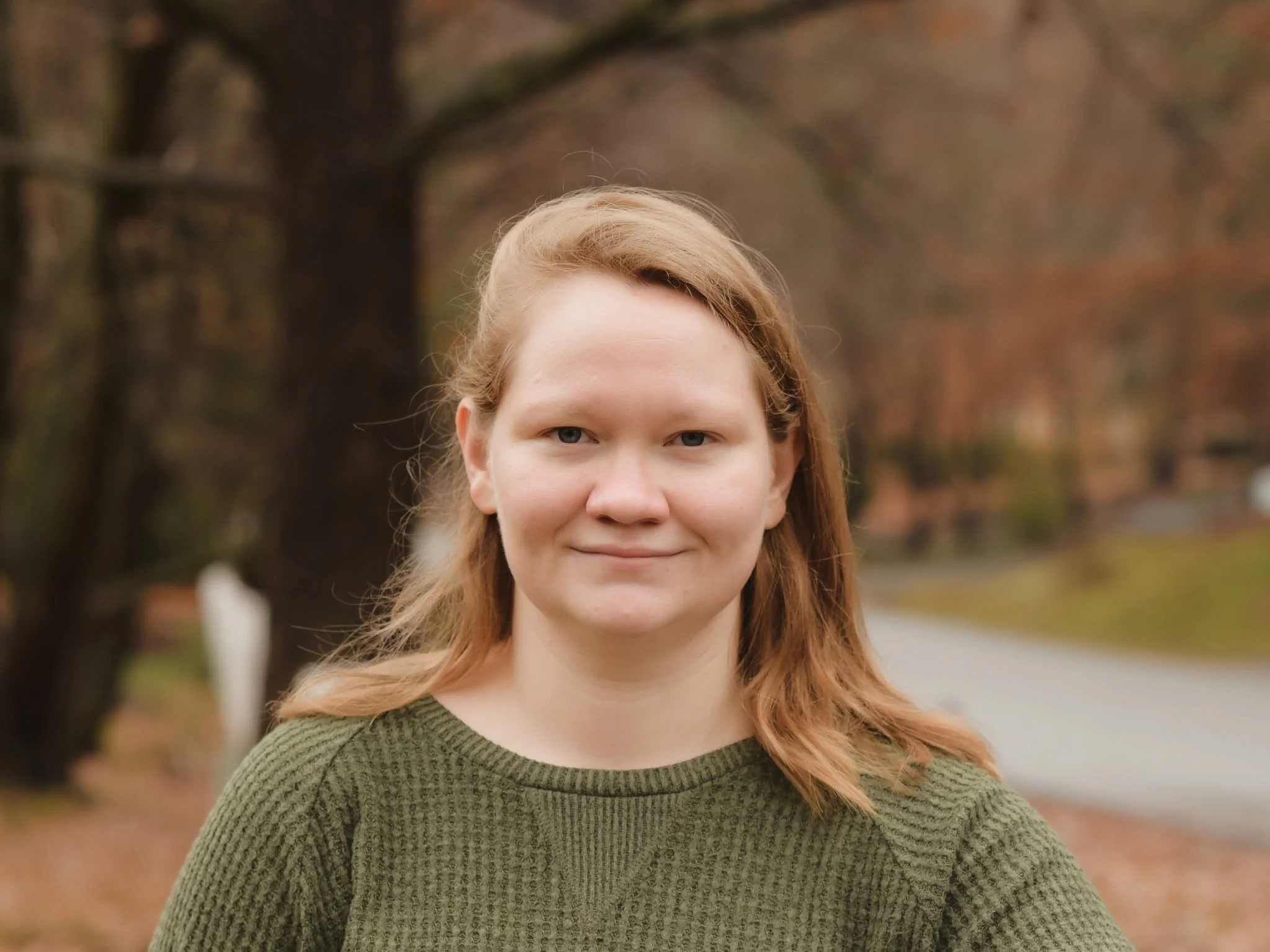 A young woman with light skin and shoulder-length blond hair standing outdoors in a park during autumn, wearing a green sweater with a shrug smile.
