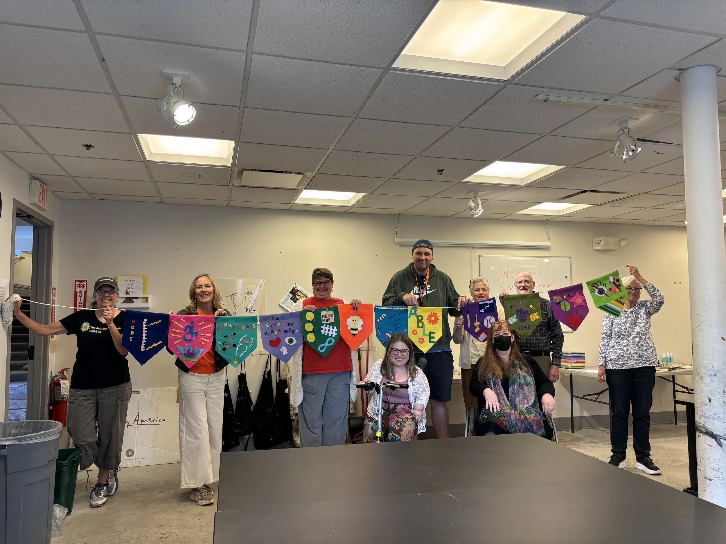 Bri sits in front of a row of people standing and holding a pennant banner made during community workshop.