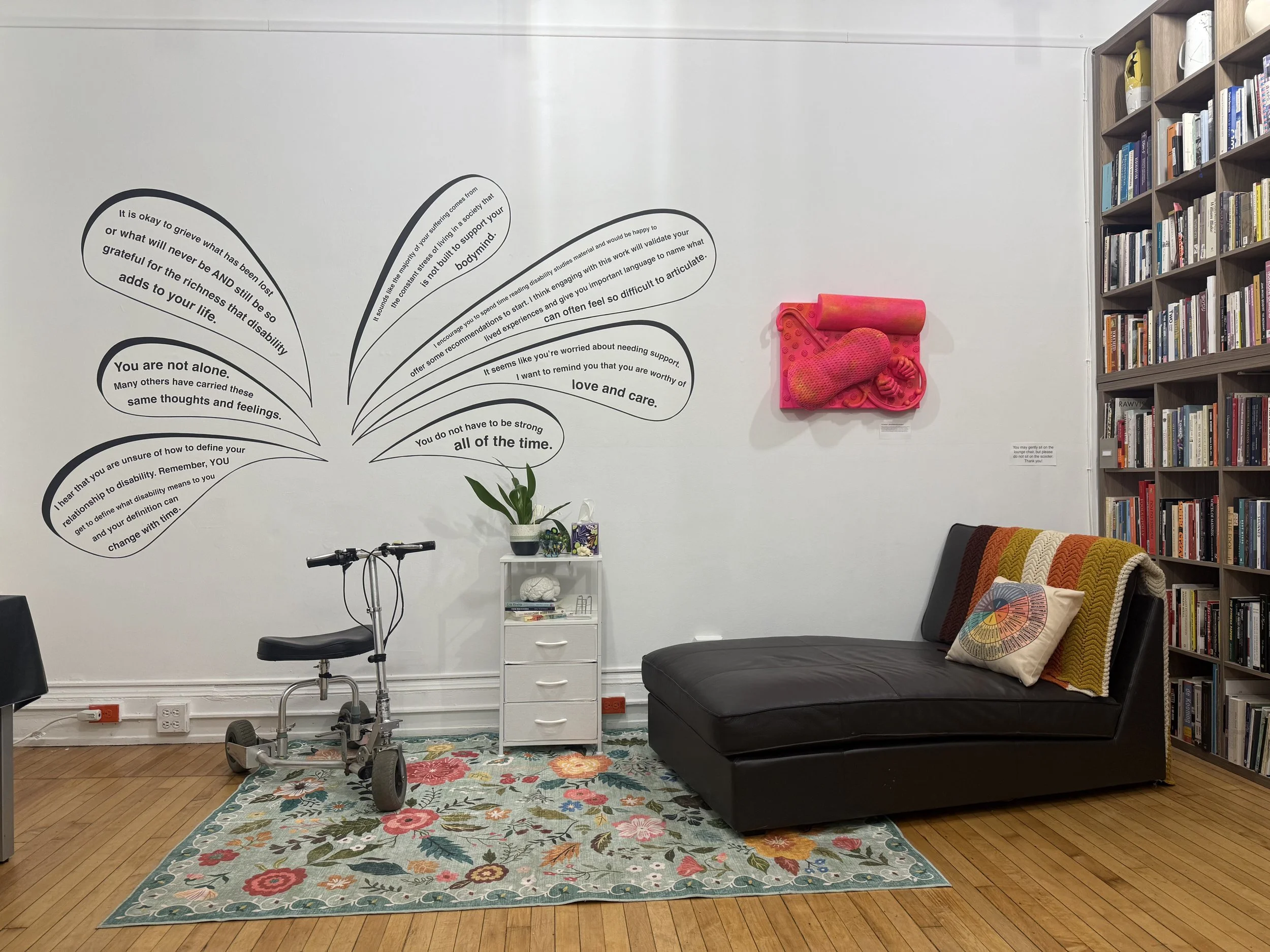 Installation display from "[From-Within]" exhibition; Bri Beck; Fall 2025; The Center for Mad Culture; Image description: A staged therapy room in an art gallery shows a mobility scooter and chase lounge facing each other on top of a floral rug. Abov