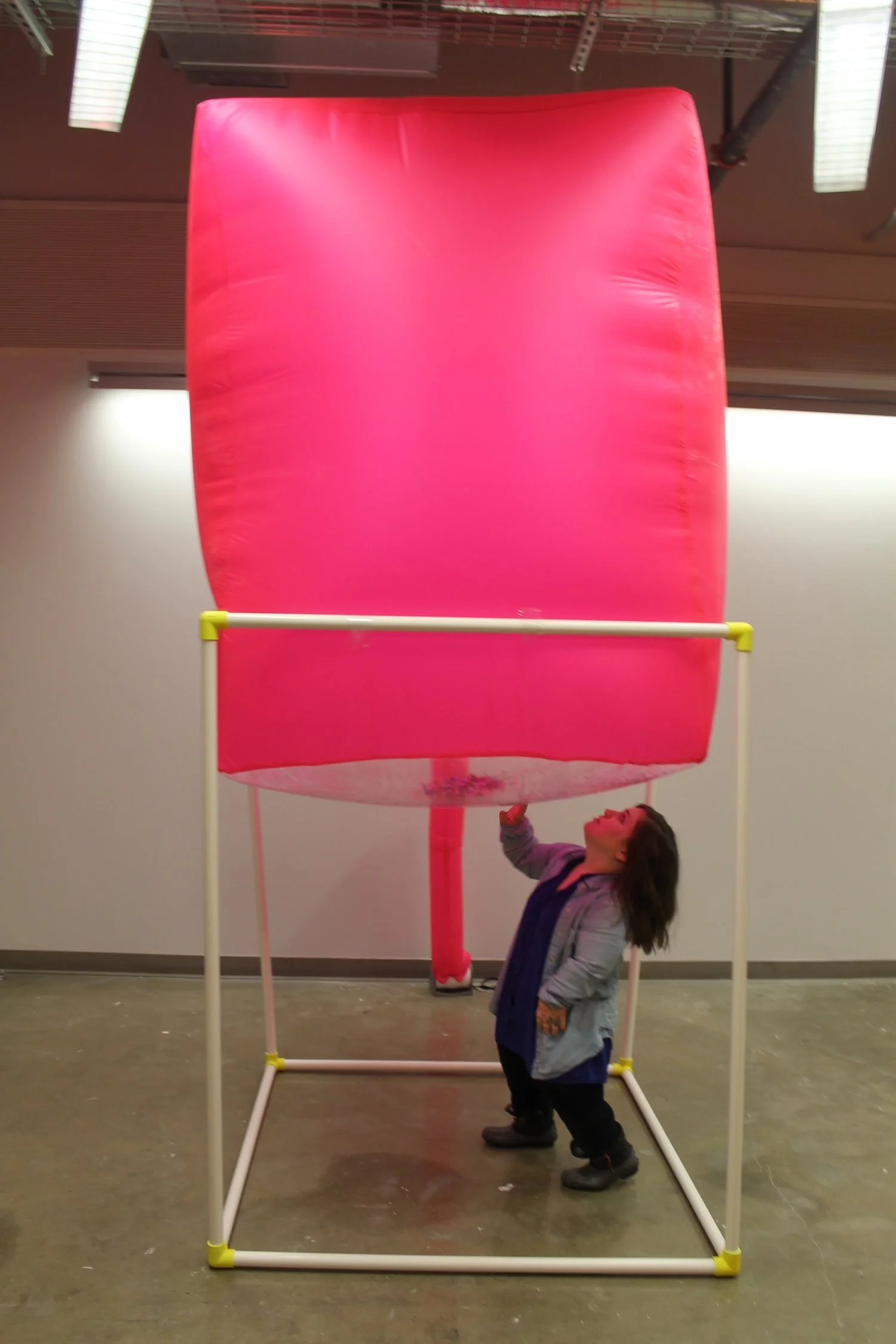 "Negotiating Space"; Installation/Interactive Sculpture; Spring 2017; Image description: Bri, a white woman of short stature, interacts with her sculpture, which is made from PVC pipe and hot pink fabric. The pvc pipe creates a rectangular prism shap