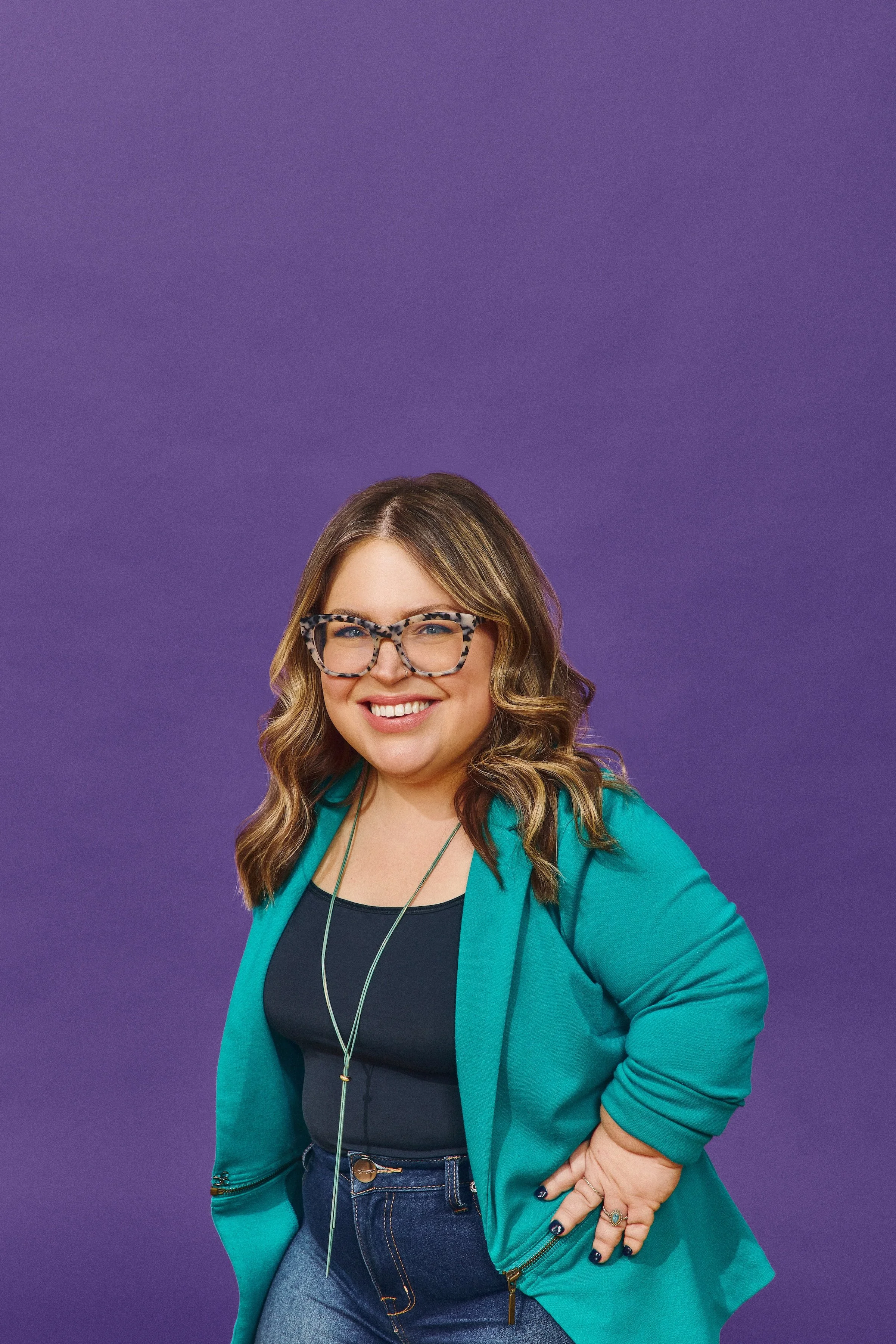 A photo of Bri, a white woman of short stature with brown wavy hair, is standing against a purple background. She is wearing a teal blazer and has her left hand on her hip.