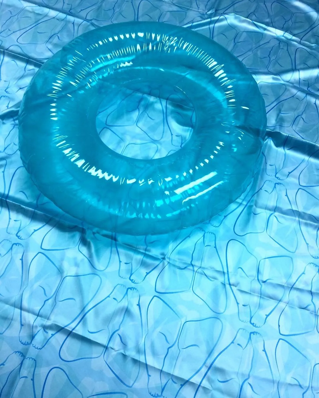 "Floating"; Photograph of Found Object on Custom Fabric; Spring 2019; Image Description: This photograph shows a clear blue inflated pool tube sitting on top of satin fabric, which resembles pool water. The design in the fabric is a repeated sketch o