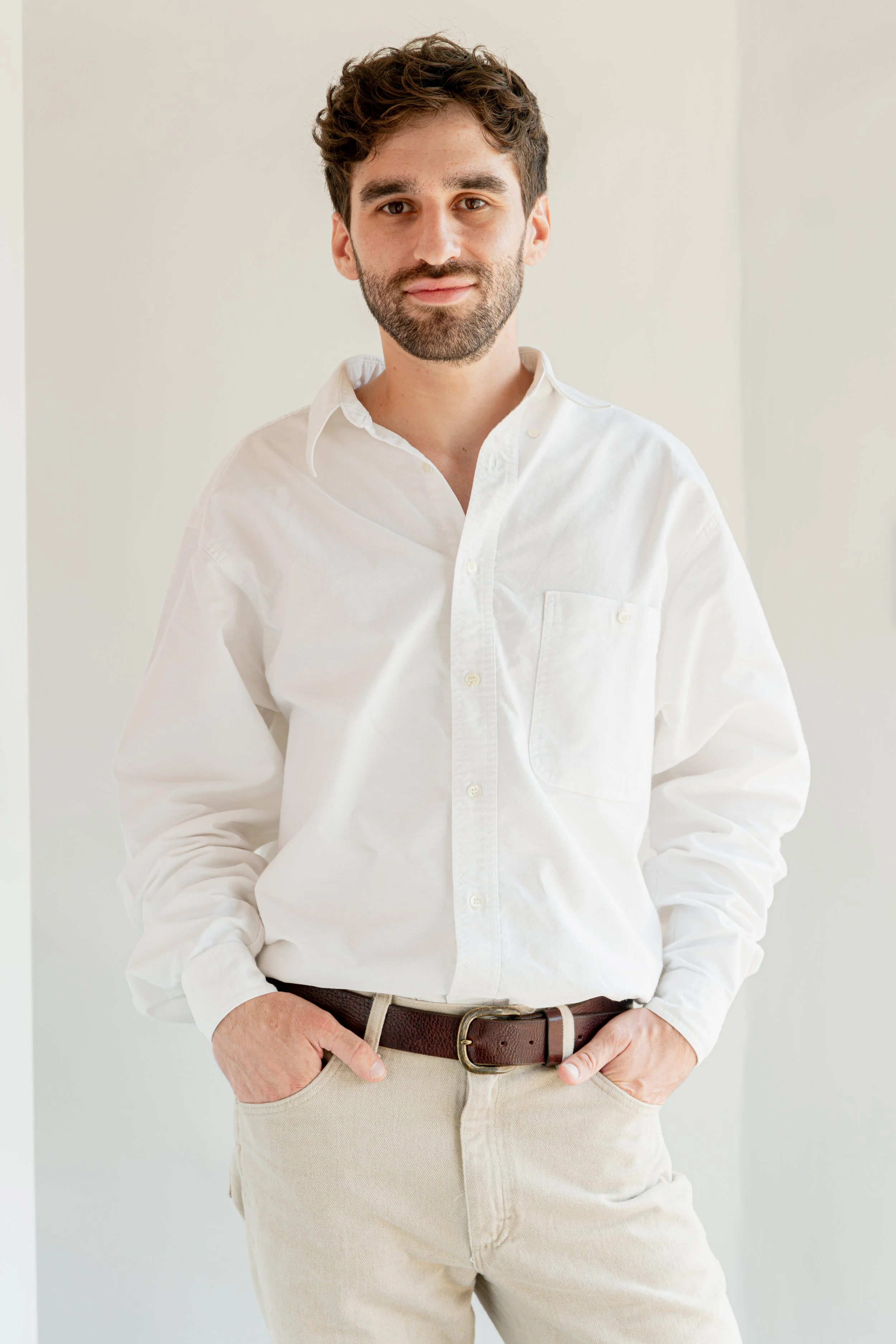 A man with brown curly hair and a beard wearing a white button-up shirt and beige pants with a brown belt standing against a plain light-colored background.