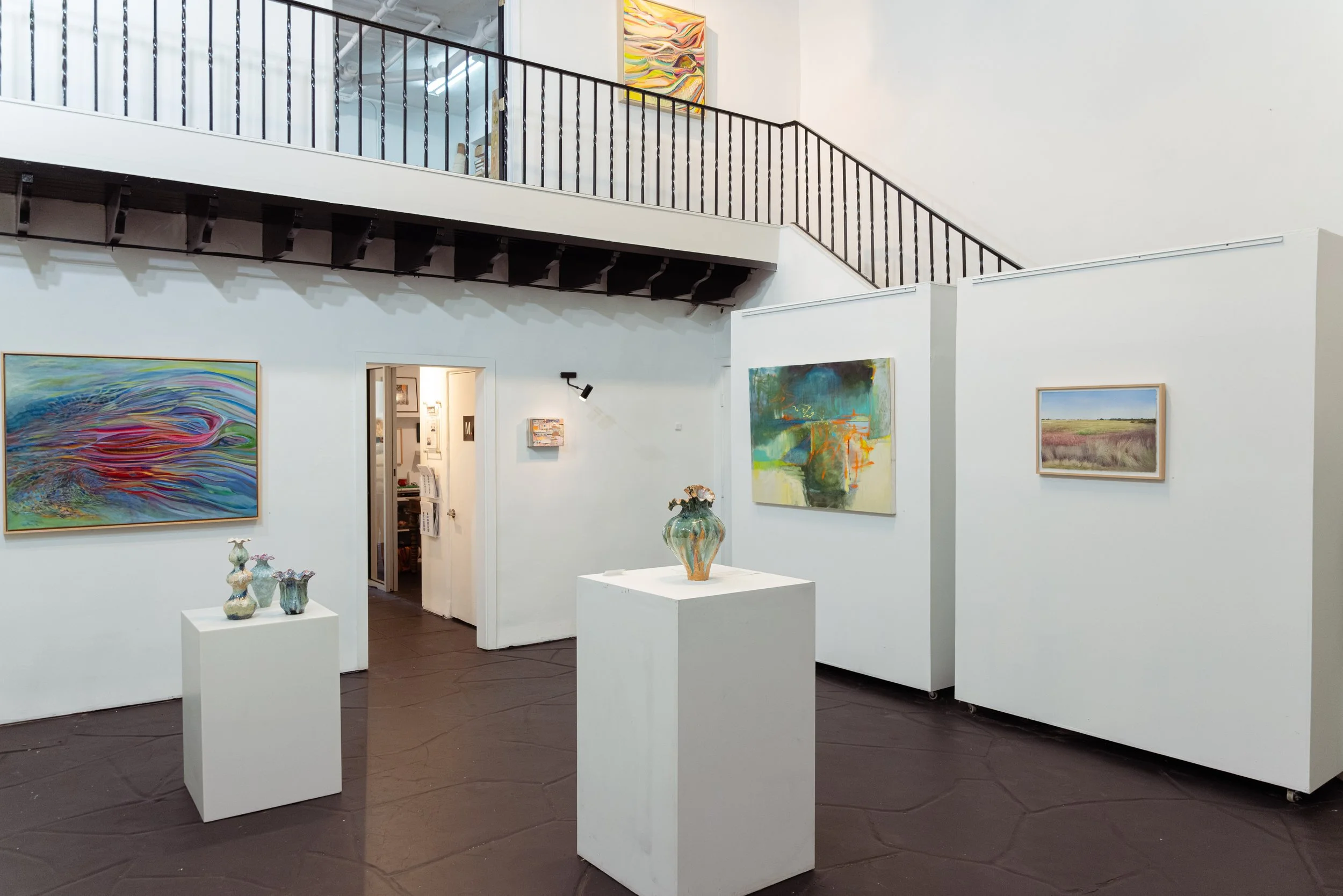 Interior of an art gallery featuring various paintings on white walls, four vases on white pedestals, and an upper balcony with a black railing.