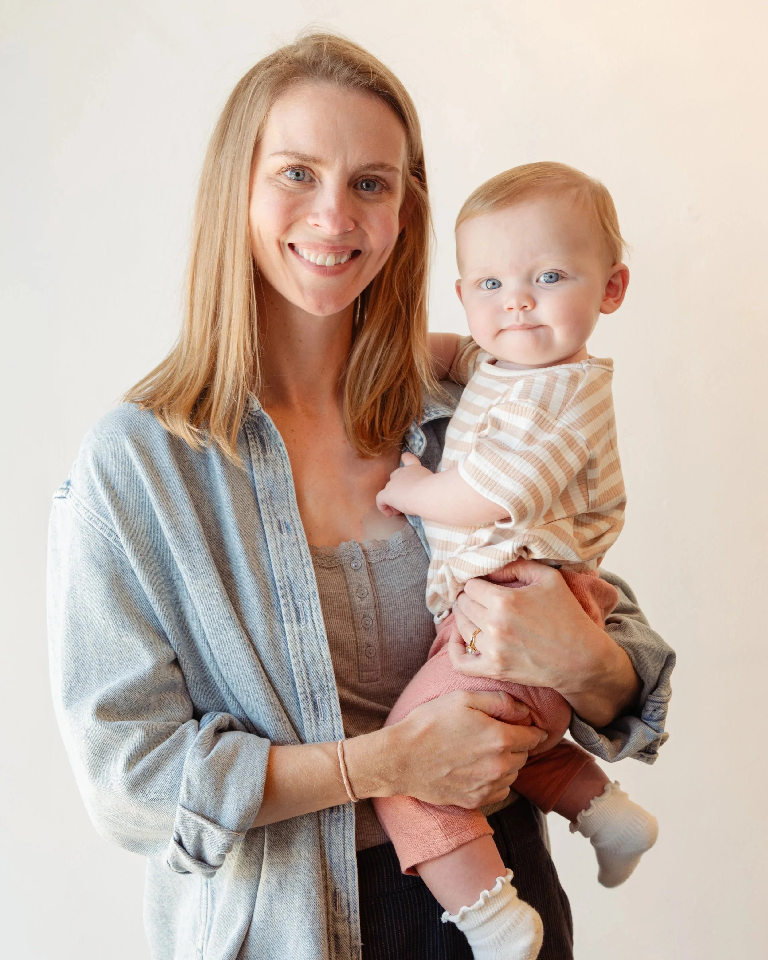 A woman with long red hair and blue eyes is smiling while holding a young child with red hair and blue eyes. The woman is wearing a denim jacket over a tan top, and the child is dressed in a striped shirt and pink shorts.