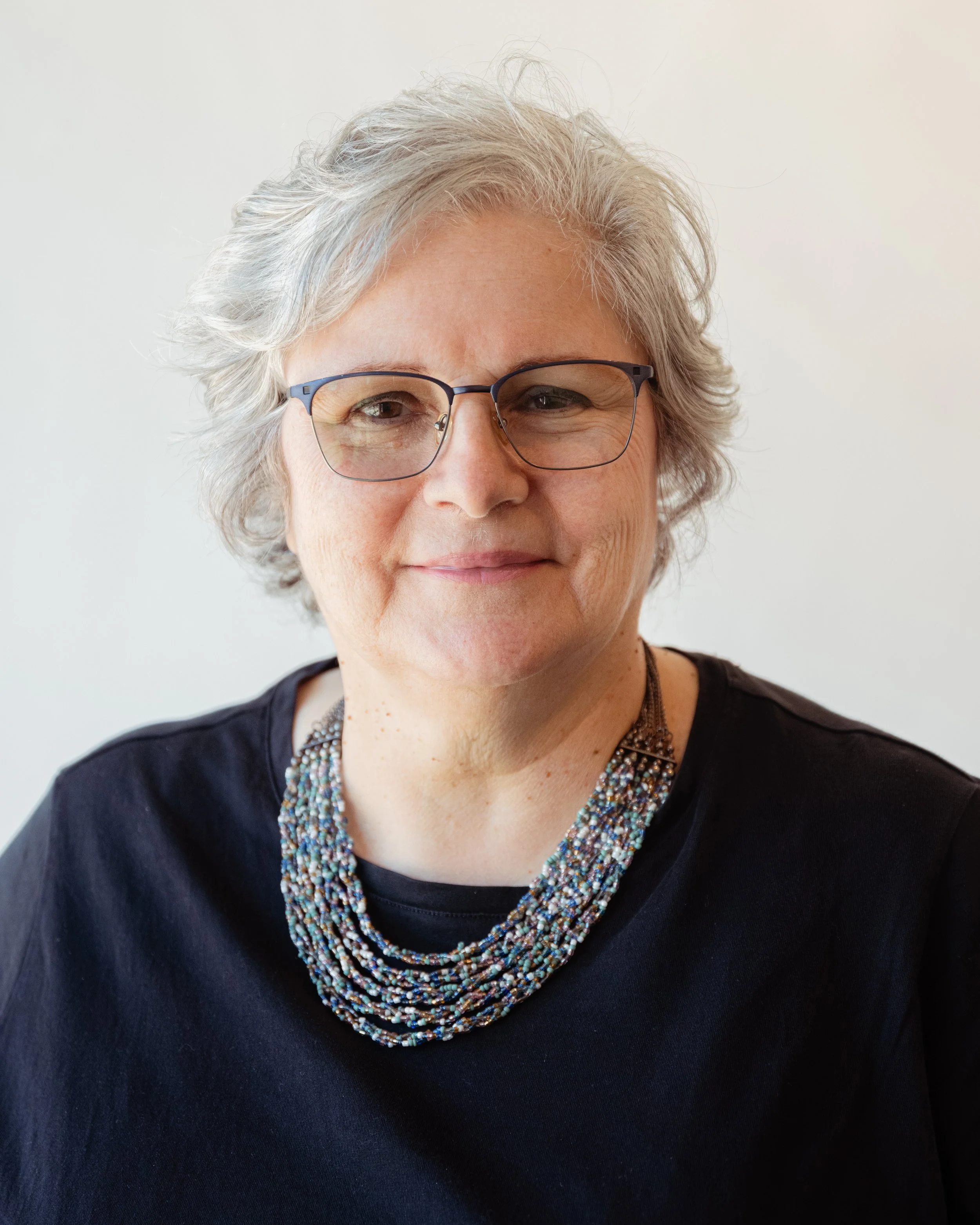 Close-up of a smiling middle-aged woman with gray hair wearing glasses and a beaded necklace, dressed in a black top, against a plain light background.
