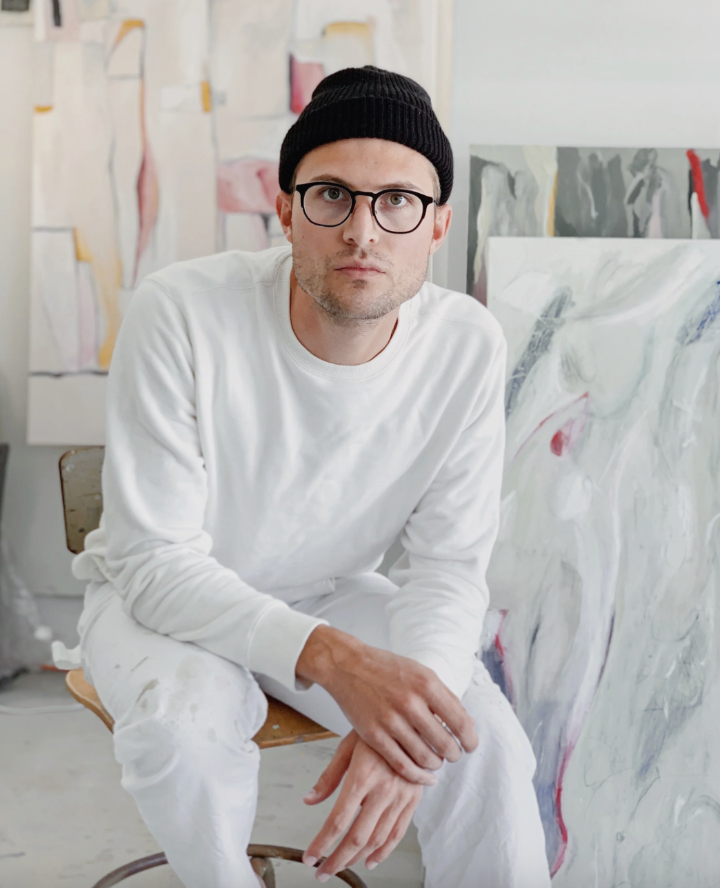 A man wearing a black beanie, glasses, and a white long-sleeve shirt sitting on a wooden chair in an art studio with abstract paintings in the background.