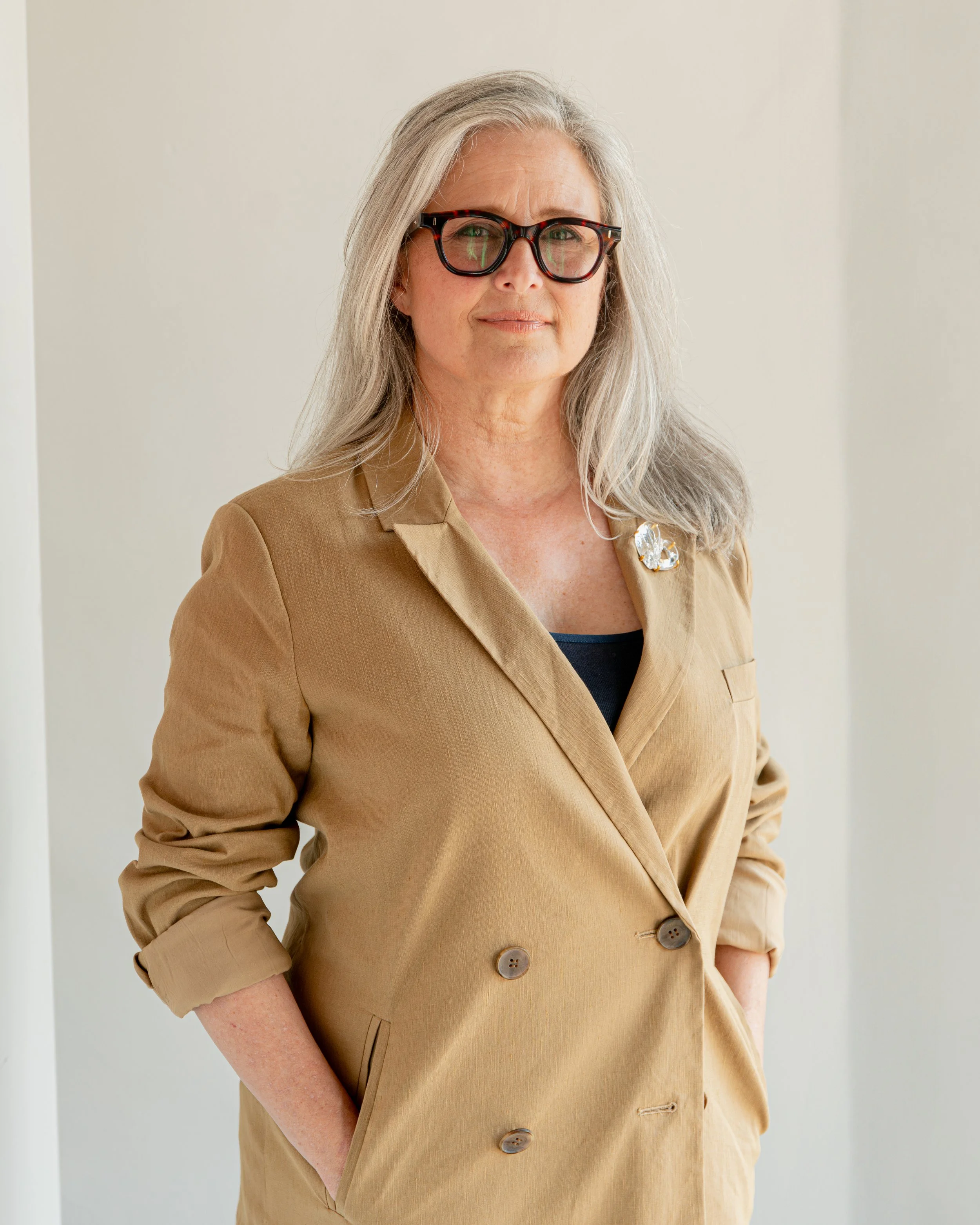 A woman with long gray hair wearing glasses with black and red frames, a tan blazer with rolled-up sleeves, and a brooch on her lapel, standing against a plain off-white wall.