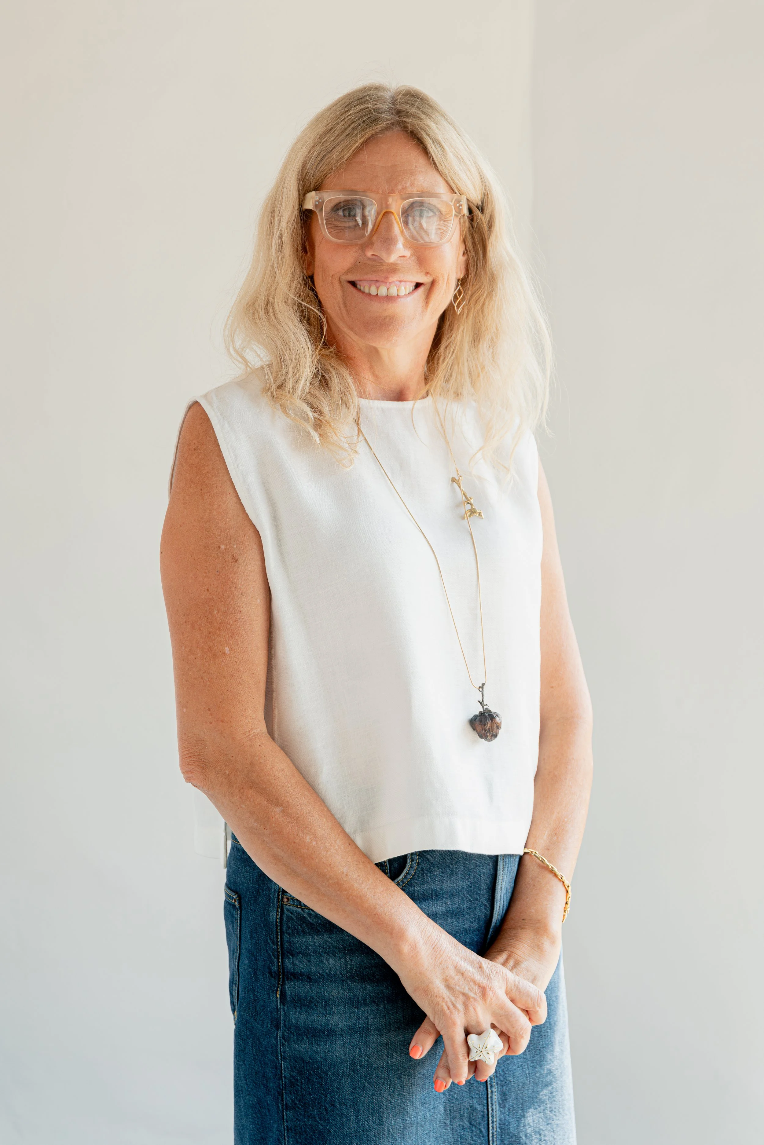 A woman with shoulder-length blonde hair and glasses smiling, wearing a sleeveless white top, blue jeans, and gold jewelry, standing against a plain light-colored background.