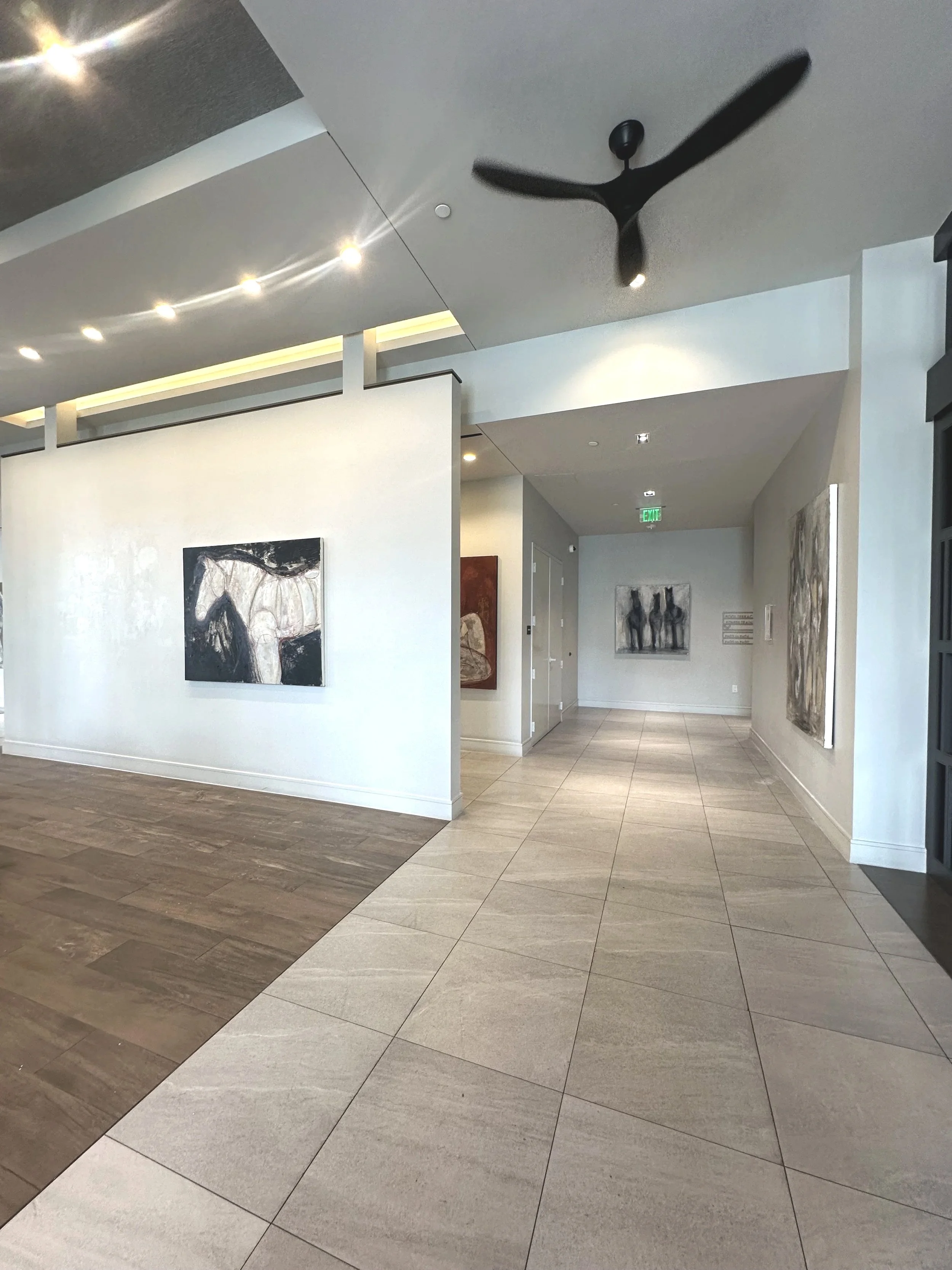 Interior of an art gallery or museum with paintings on white walls, a tile floor, and a ceiling with recessed lights and a black ceiling fan.
