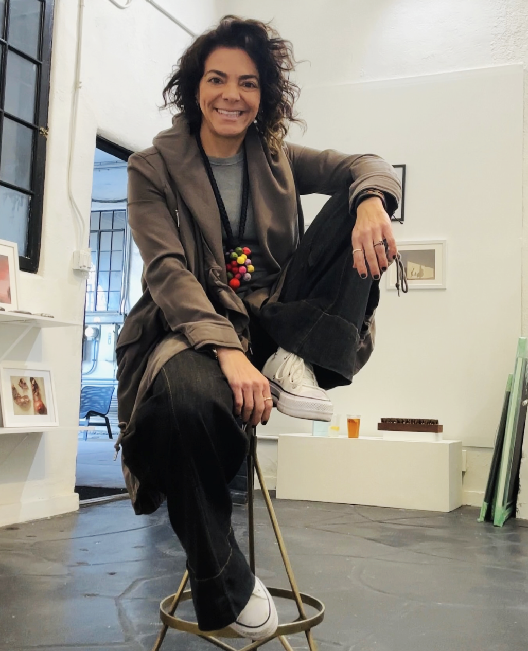 Woman with curly hair smiling, wearing a gray jacket, black pants, and white sneakers, balancing on a tall stool indoors.