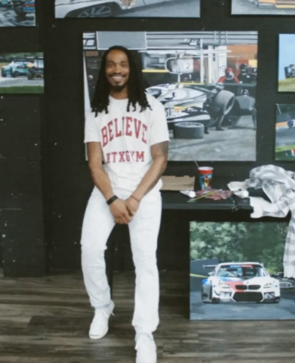 A man with dreadlocks smiling and standing indoors in front of a display of racing and sports cars, with artwork and photos on the wall behind him.