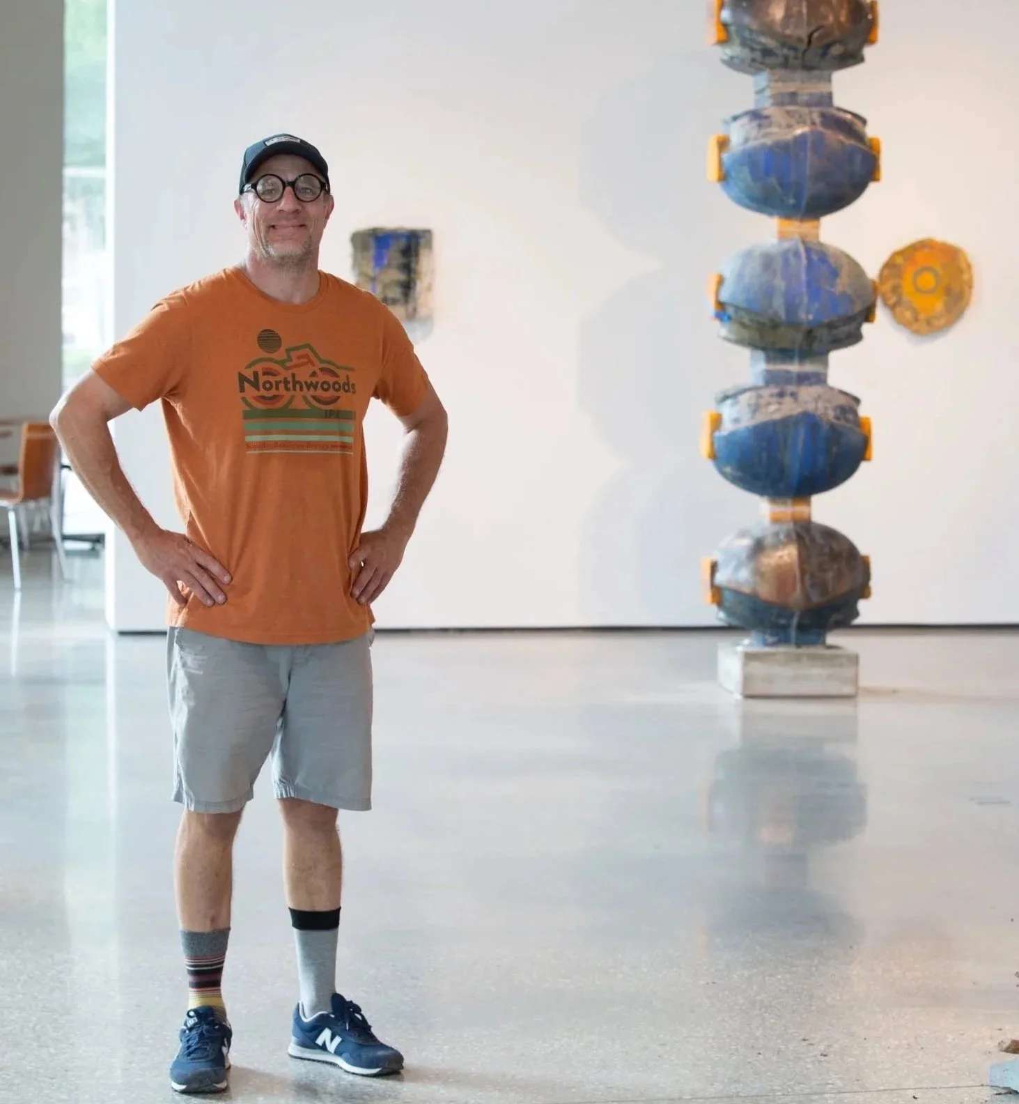 A man with glasses, wearing an orange T-shirt with 'Northwoods' written on it, khaki shorts, and sneakers, stands in an art gallery with colorful abstract sculptures in the background.