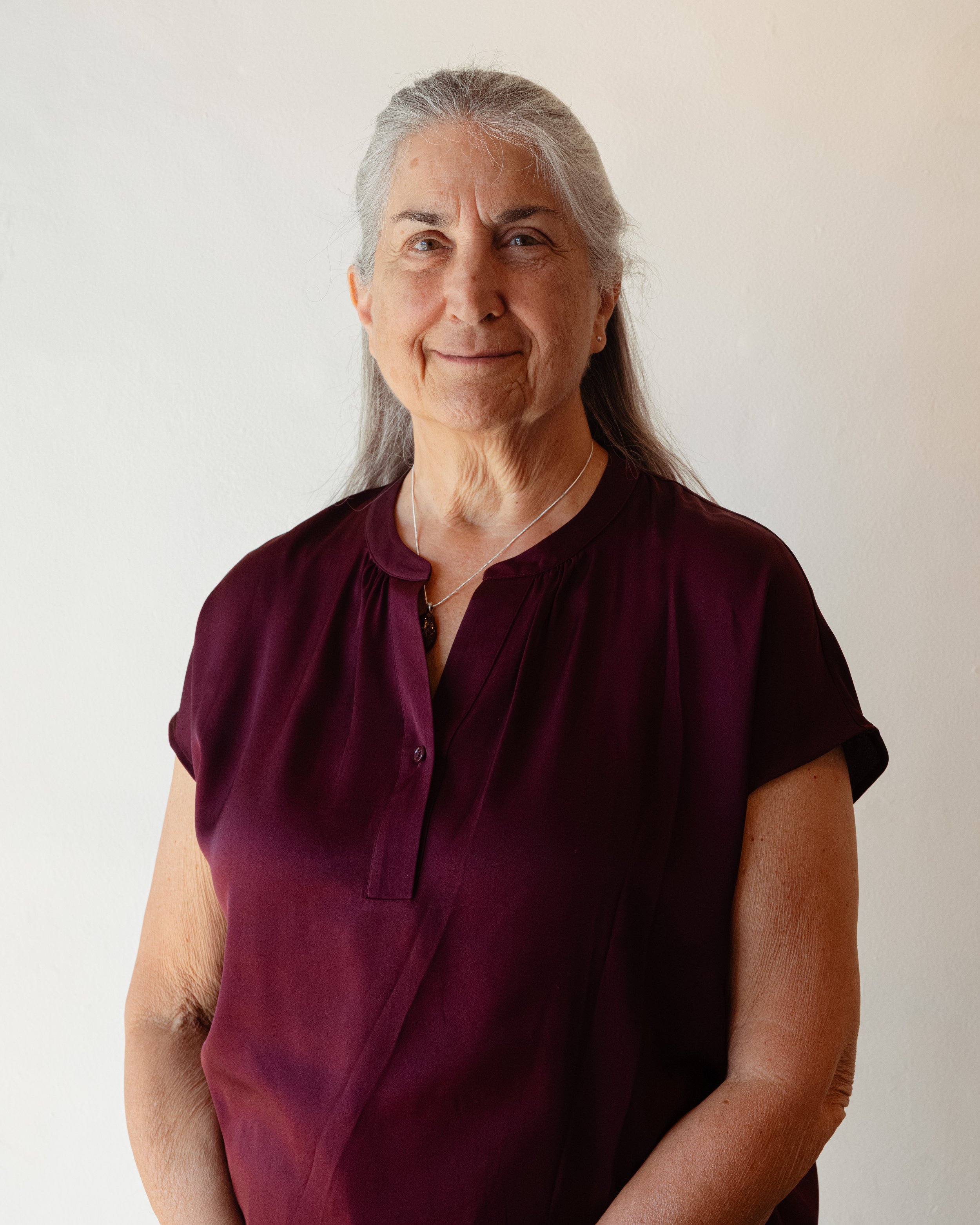Close-up of a middle-aged woman with gray hair, wearing a maroon blouse and a silver necklace, smiling gently at the camera against a plain white background.