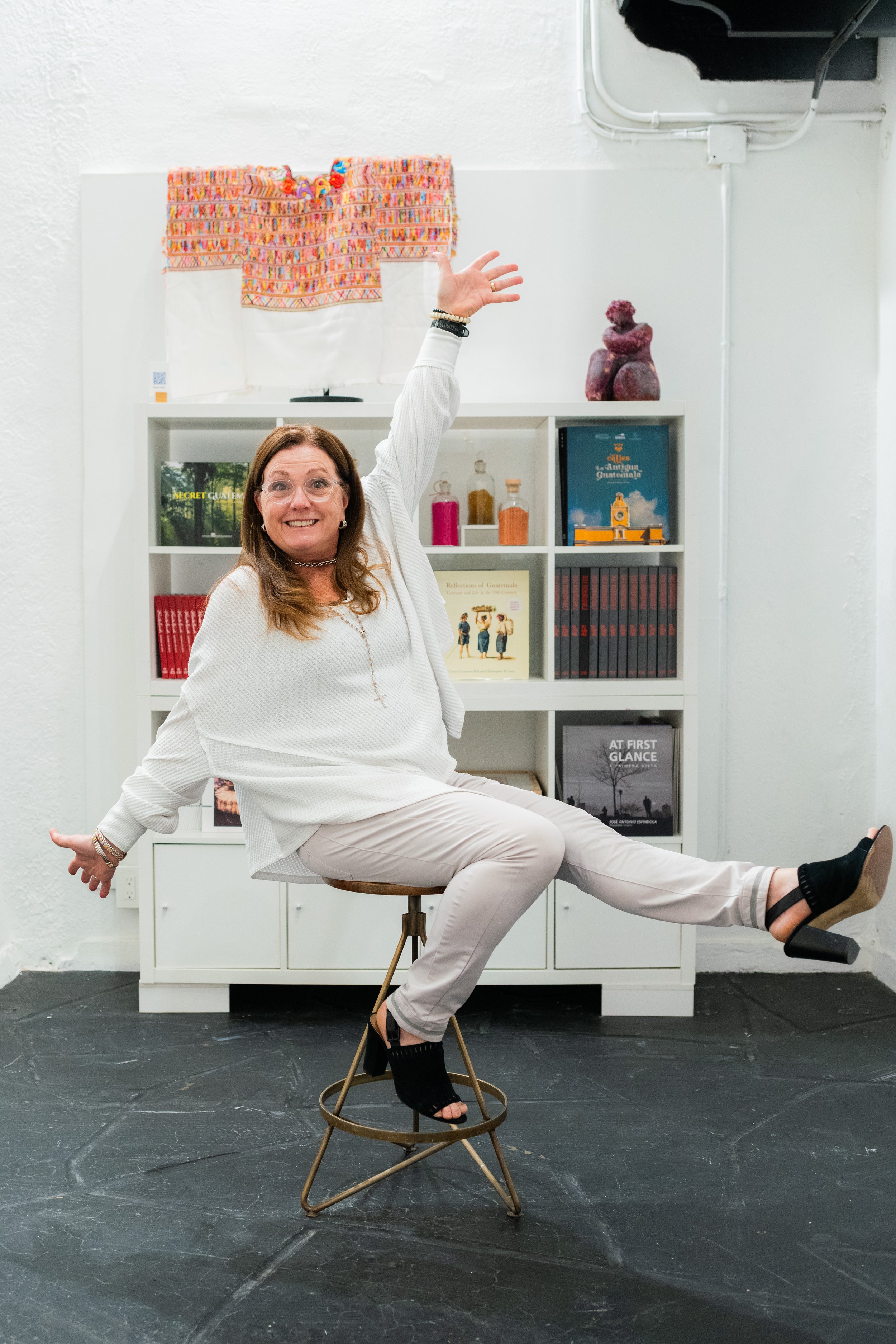 A woman with glasses and brown hair smiling, sitting on a stool and posing with her right arm raised and her left arm extended to the side. She is wearing a white long-sleeve top and light-colored pants, with one leg crossed over the other. Behind her is a white bookshelf with books and bottles, with a colorful embroidered textile hanging above and a small sculpture on top.