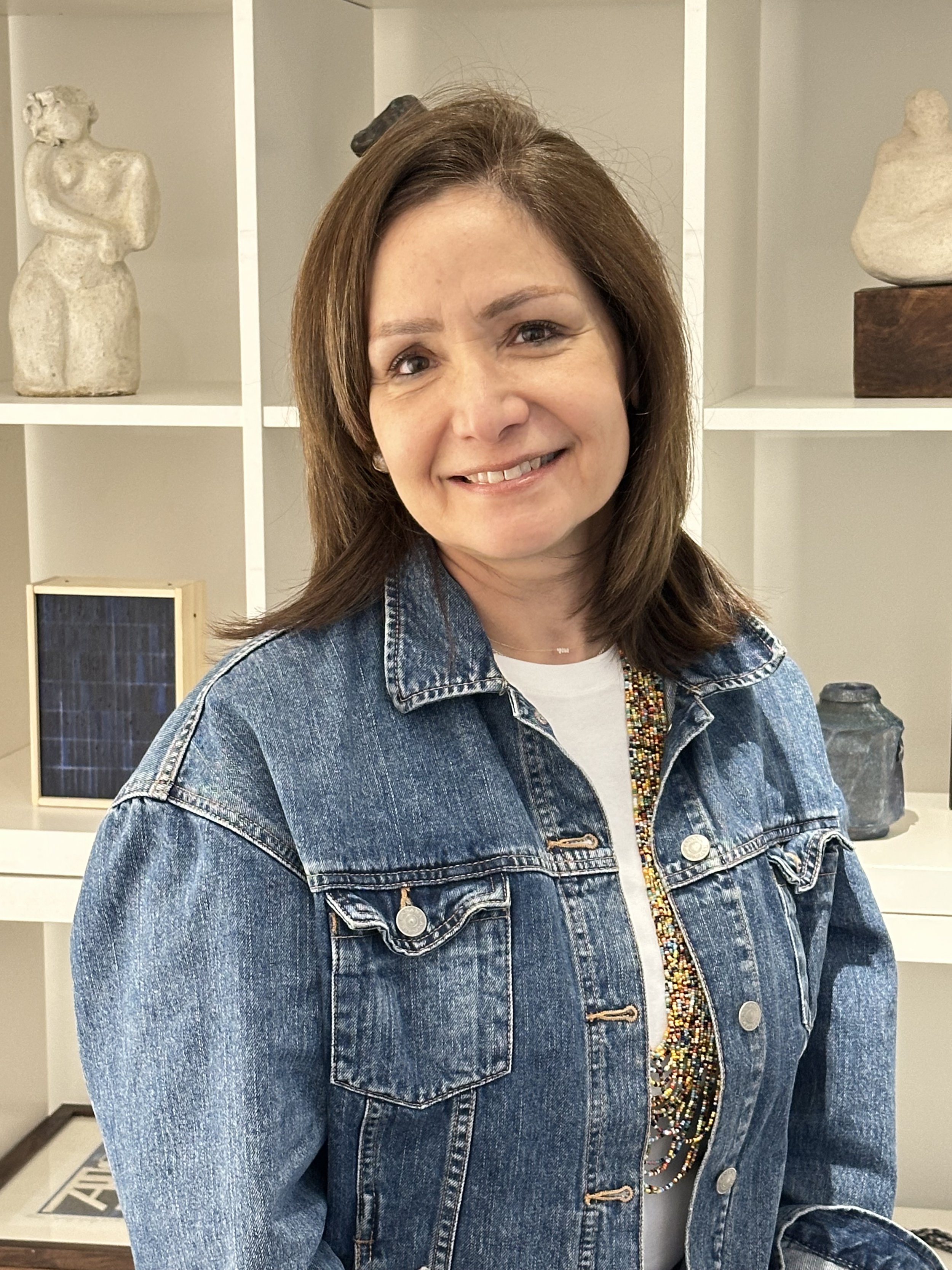 A woman with brown hair smiling, wearing a denim jacket and a multi-colored beaded necklace, standing in front of a white shelf with decorative sculptures.