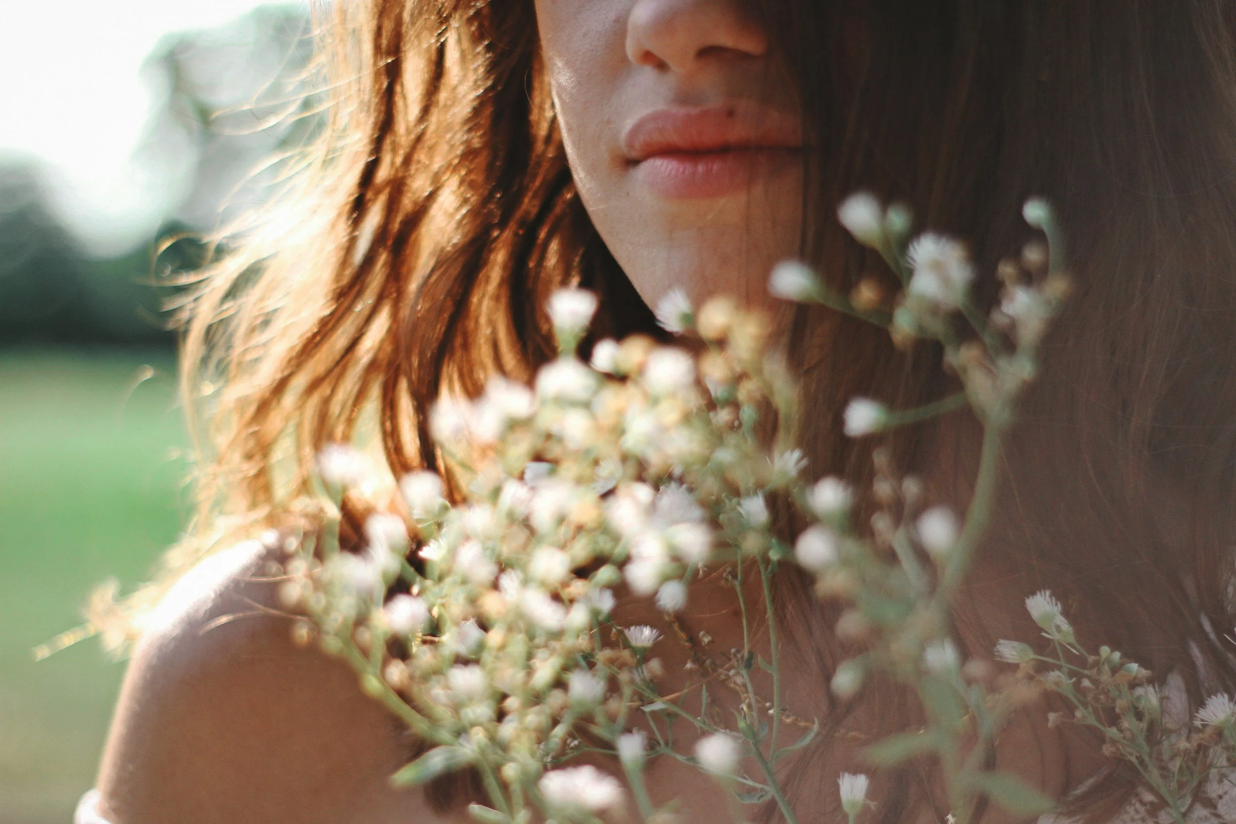 A woman with red hair holding white flowers close to her face outdoors during daytime.