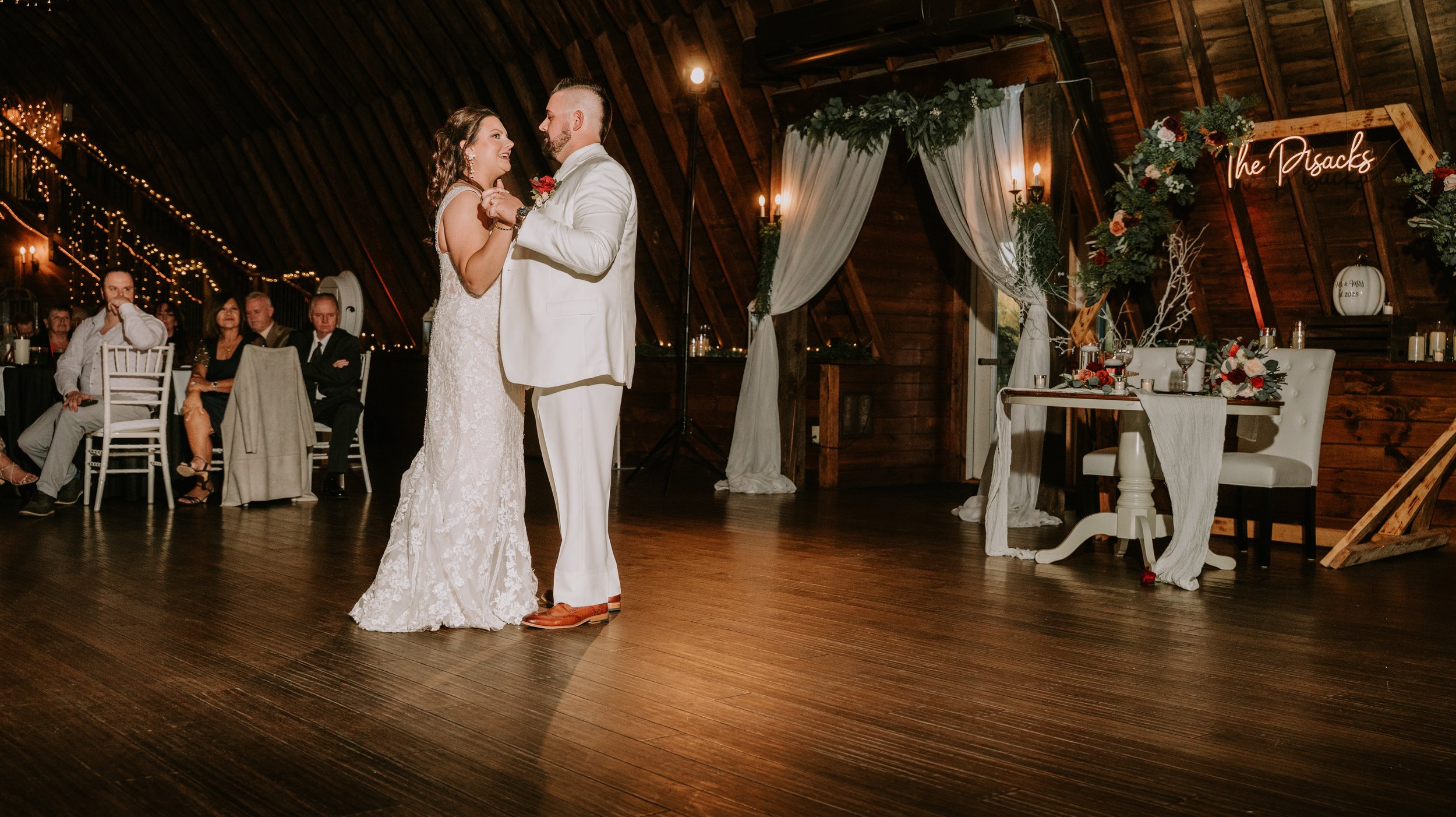 A bride and groom dance during their wedding reception in a rustic wooden venue with fairy lights, seated guests watching, and wedding decor including a floral arch, a table with flowers and candles, and a neon sign that says 'The Pósacks.'