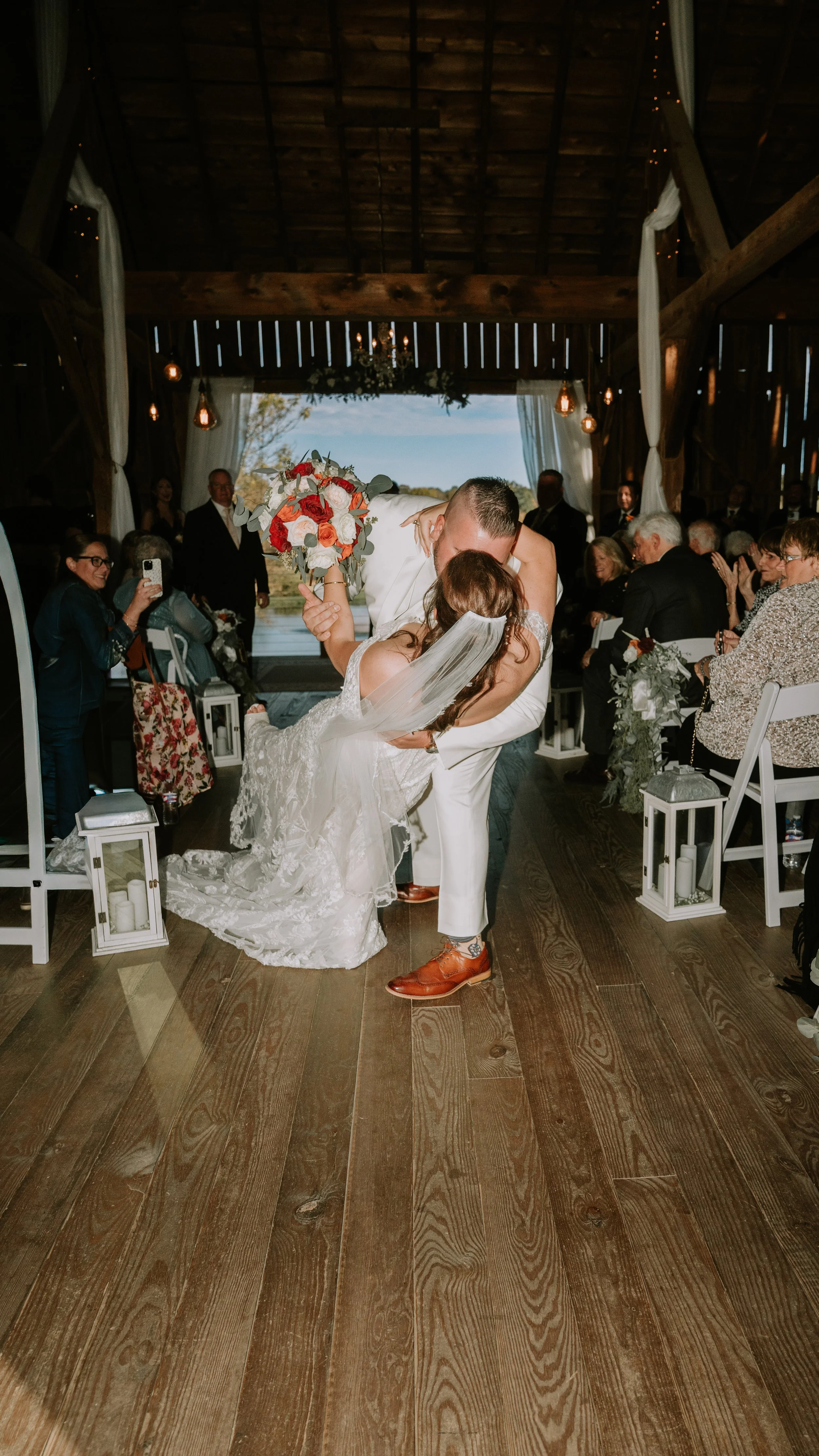 A bride and groom are kissing during their wedding ceremony inside a rustic wooden barn with guests seated around them.