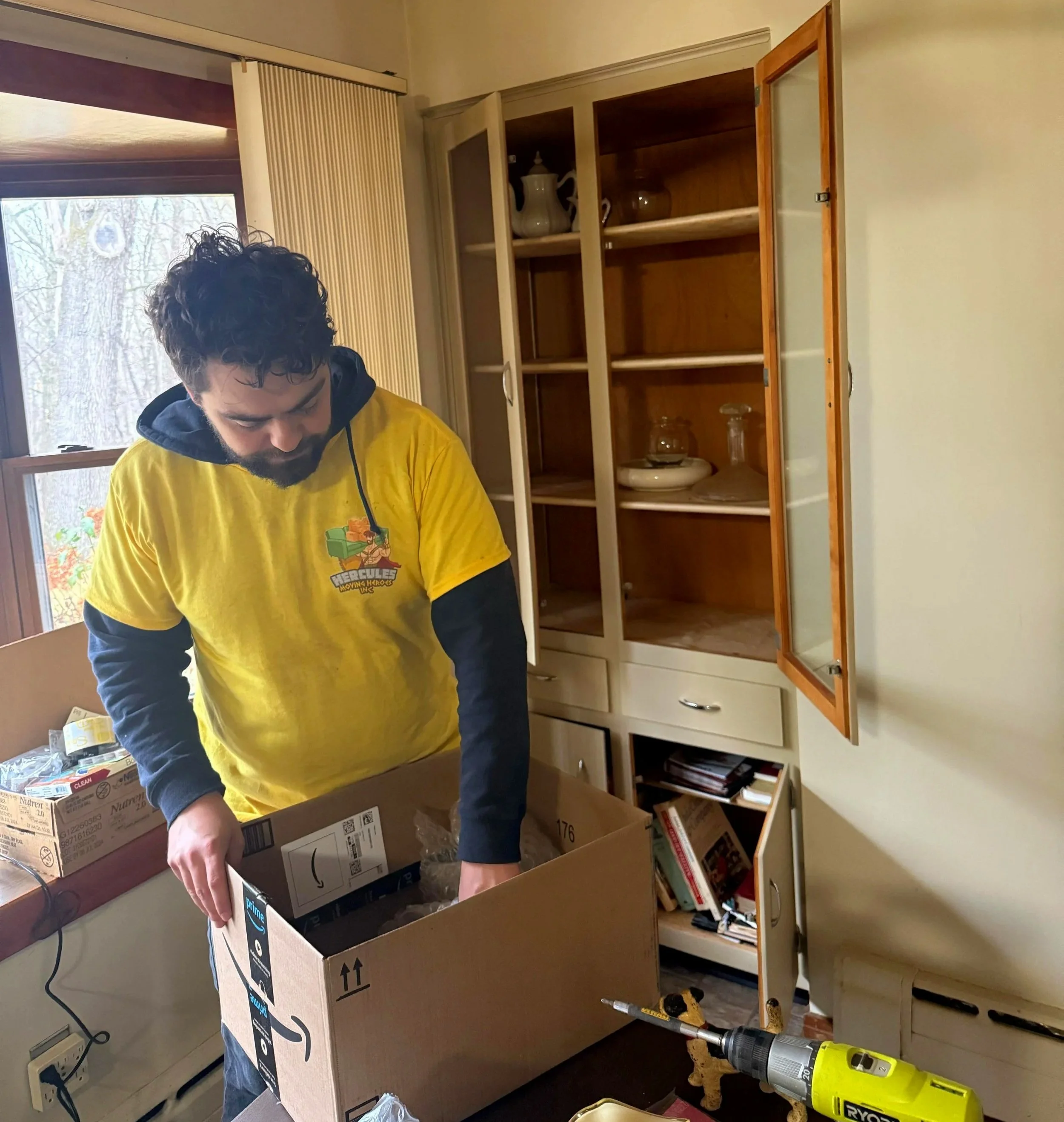 A man with curly hair and a beard in a yellow T-shirt unpacking items from a cardboard box in a room with a cabinet, window, and a workbench.