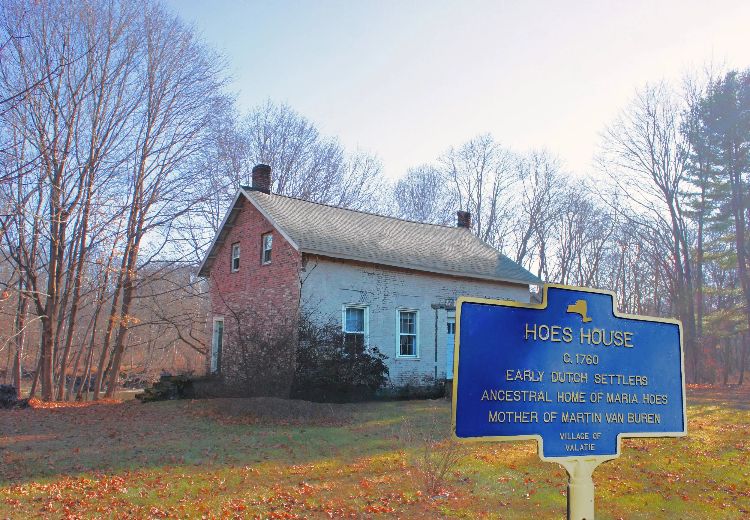 A historical marker sign in front of a house with a gray roof and brick chimney, surrounded by trees with autumn leaves on the ground, indicating the site is the early Dutch settlement and ancestral home of Maria Hoes.