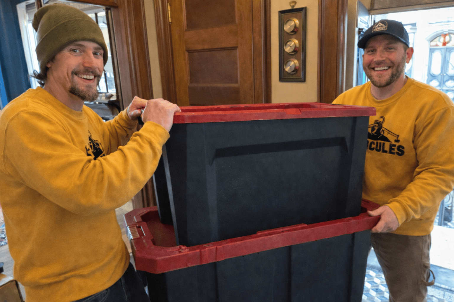 Two men smiling and holding a large black plastic storage bin with a red lid between them in a cozy, wood-paneled room.