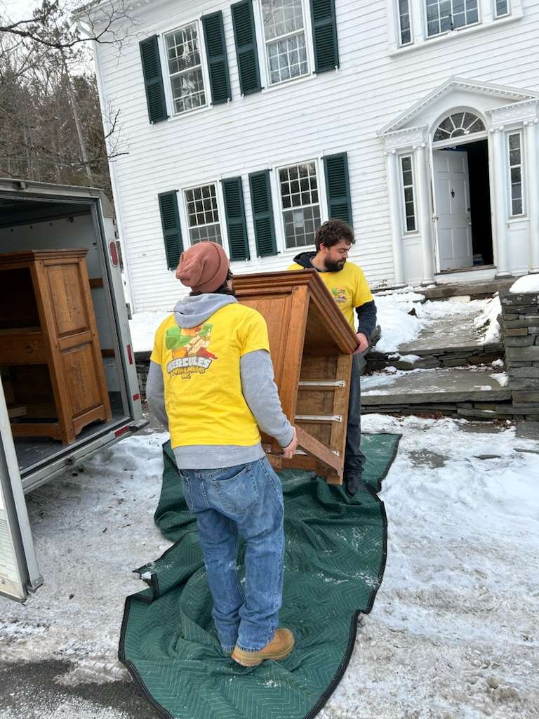 A man standing next to a white van with a red hand truck in his hand, outdoors on a sunny day with leafless trees in the background.