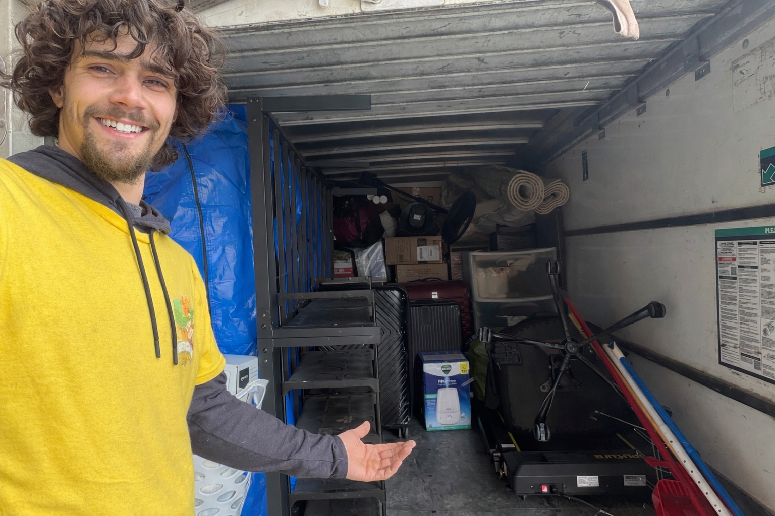 A man standing next to a white van with a red hand truck in his hand, outdoors on a sunny day with leafless trees in the background.