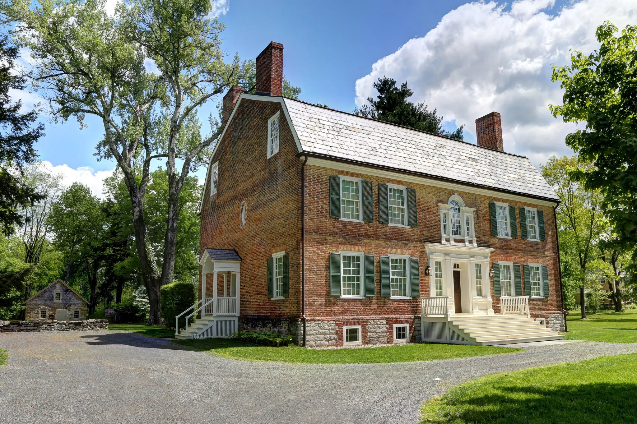 A large brick house with green shutters and a white porch, surrounded by trees under a blue sky with some clouds.