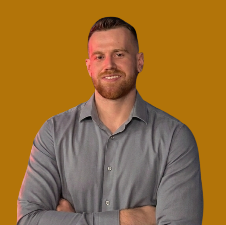 A man with a beard and short hair, wearing a gray button-up shirt, is standing with arms crossed against a brown background.