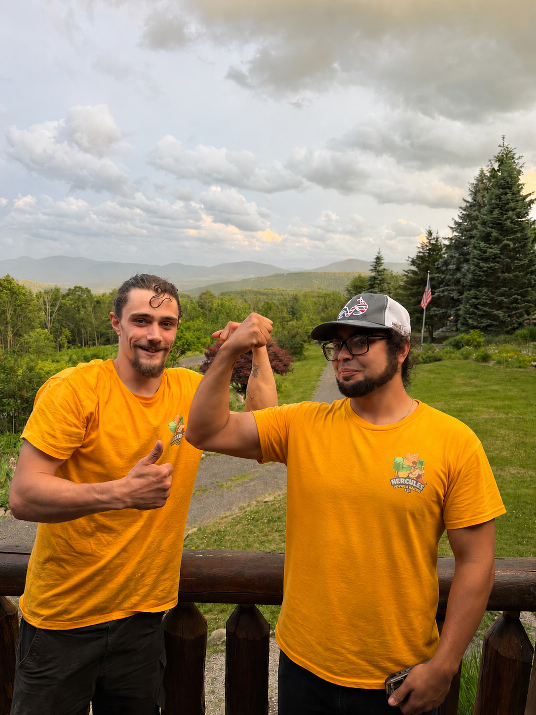 Two men in yellow t-shirts showing a strong arm pose in an outdoor setting with lush green trees, a mountain range, and a cloudy sky in the background.
