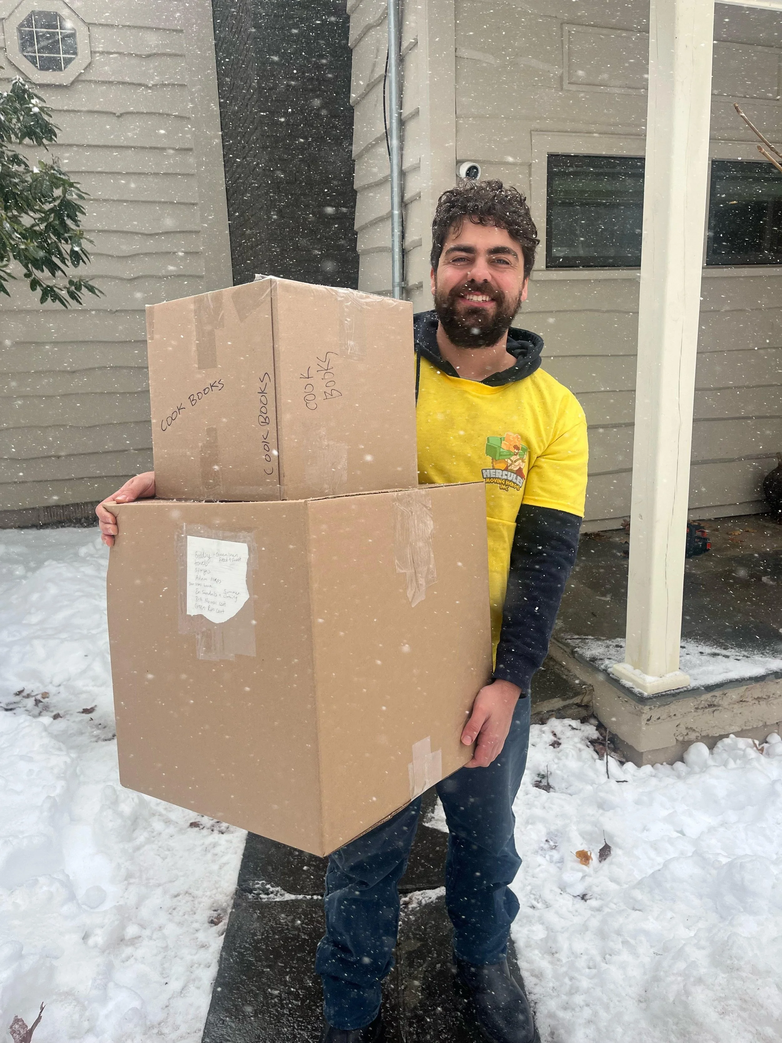 A man smiling and holding two large cardboard boxes labeled "Cook Books" stands outside in the snow, next to a house with gray siding and a porch.