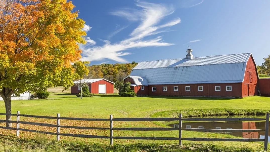 Scenic view of a red barn and smaller outbuilding on a farm with a pond, green grass, trees with fall foliage, and a blue sky with clouds.
