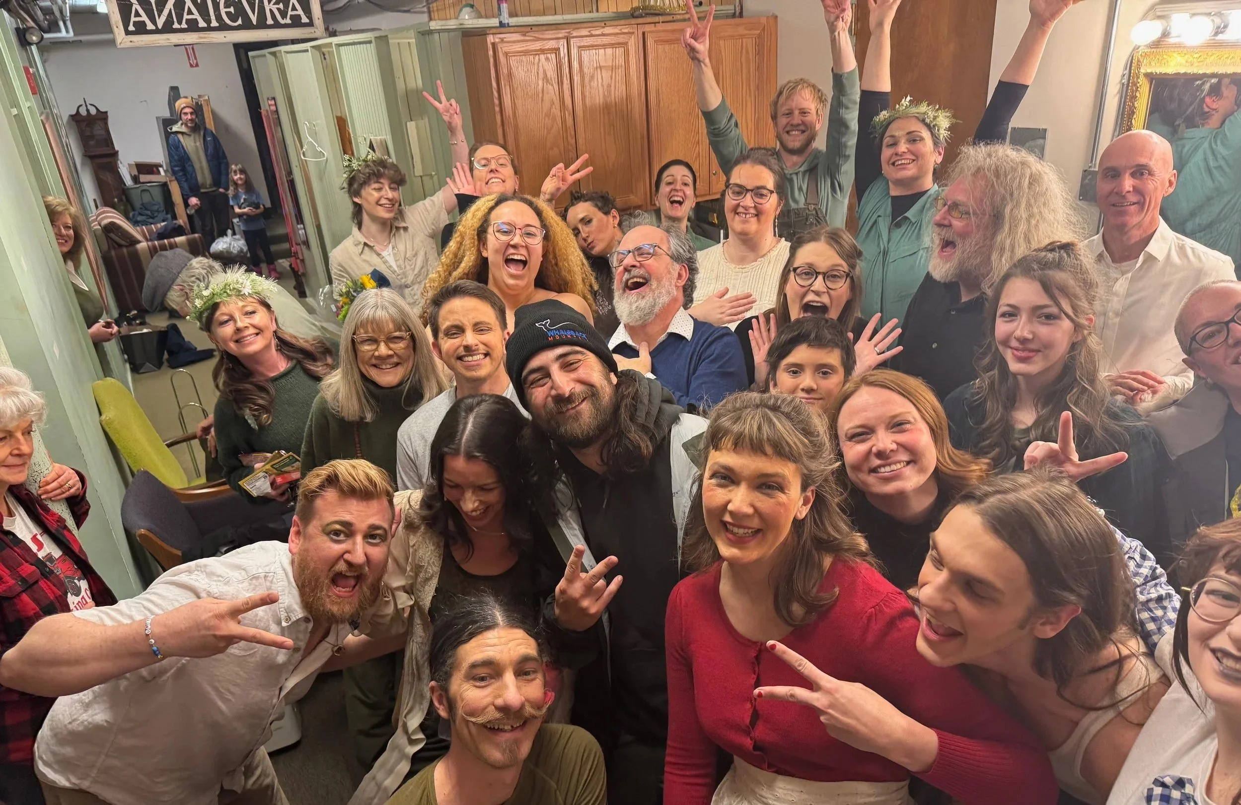 A large group of smiling people gathered indoors, celebrating, with some making peace signs and others wearing flower crowns.