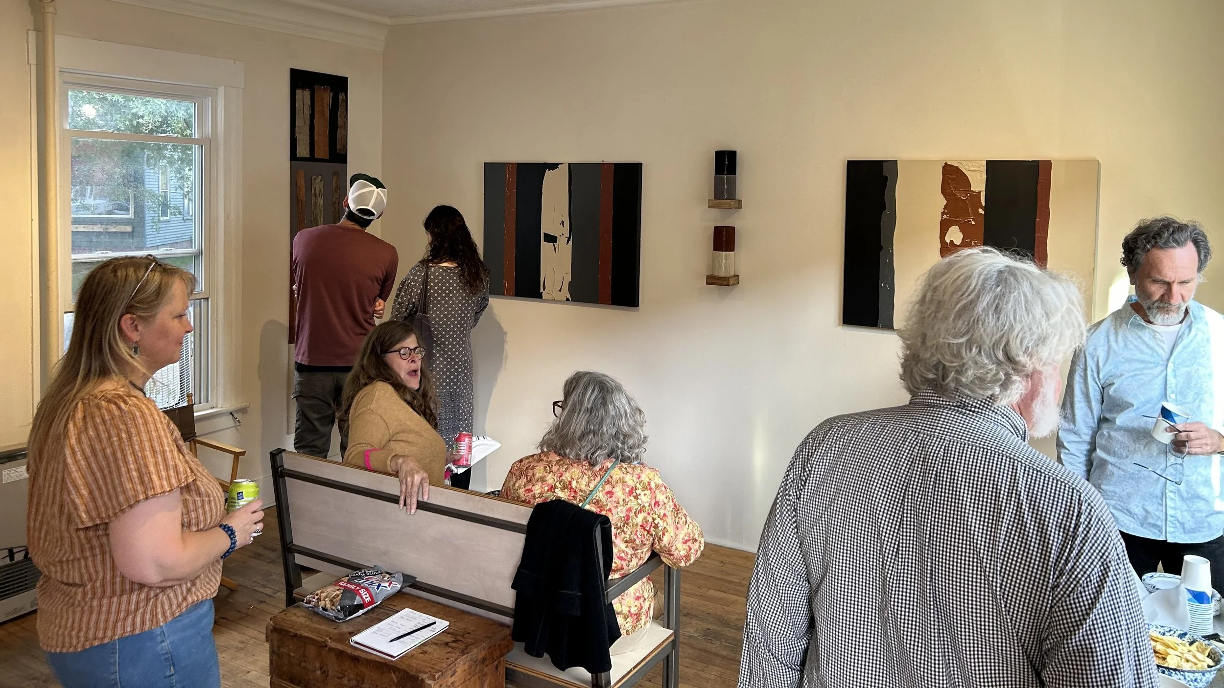 A group of people socializing in an art gallery, with several abstract paintings on the wall, some sitting, some standing, holding drinks, and chatting.