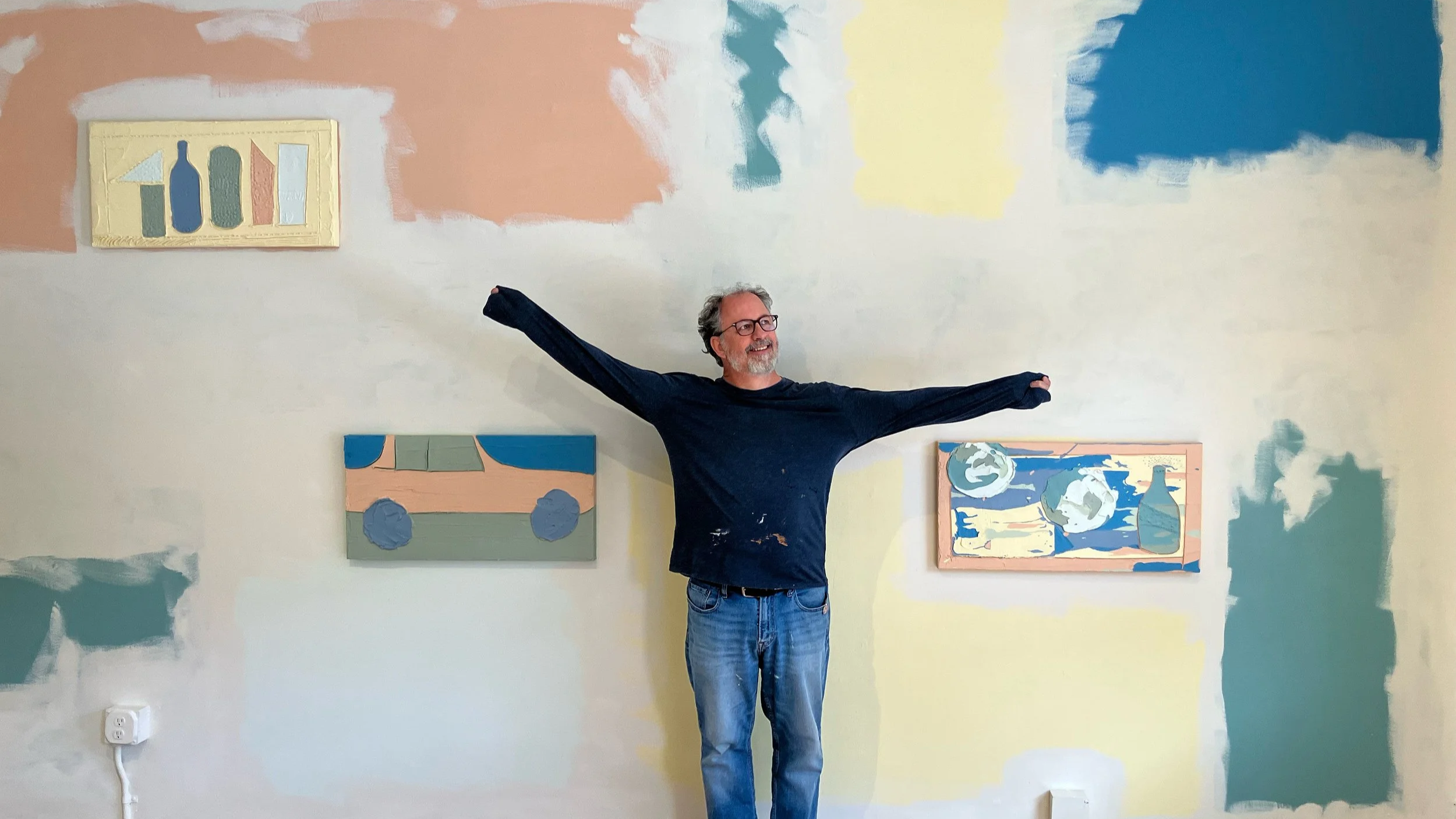 A man with gray hair, beard, and glasses standing in front of a wall with abstract paintings, stretching his arms wide and smiling.