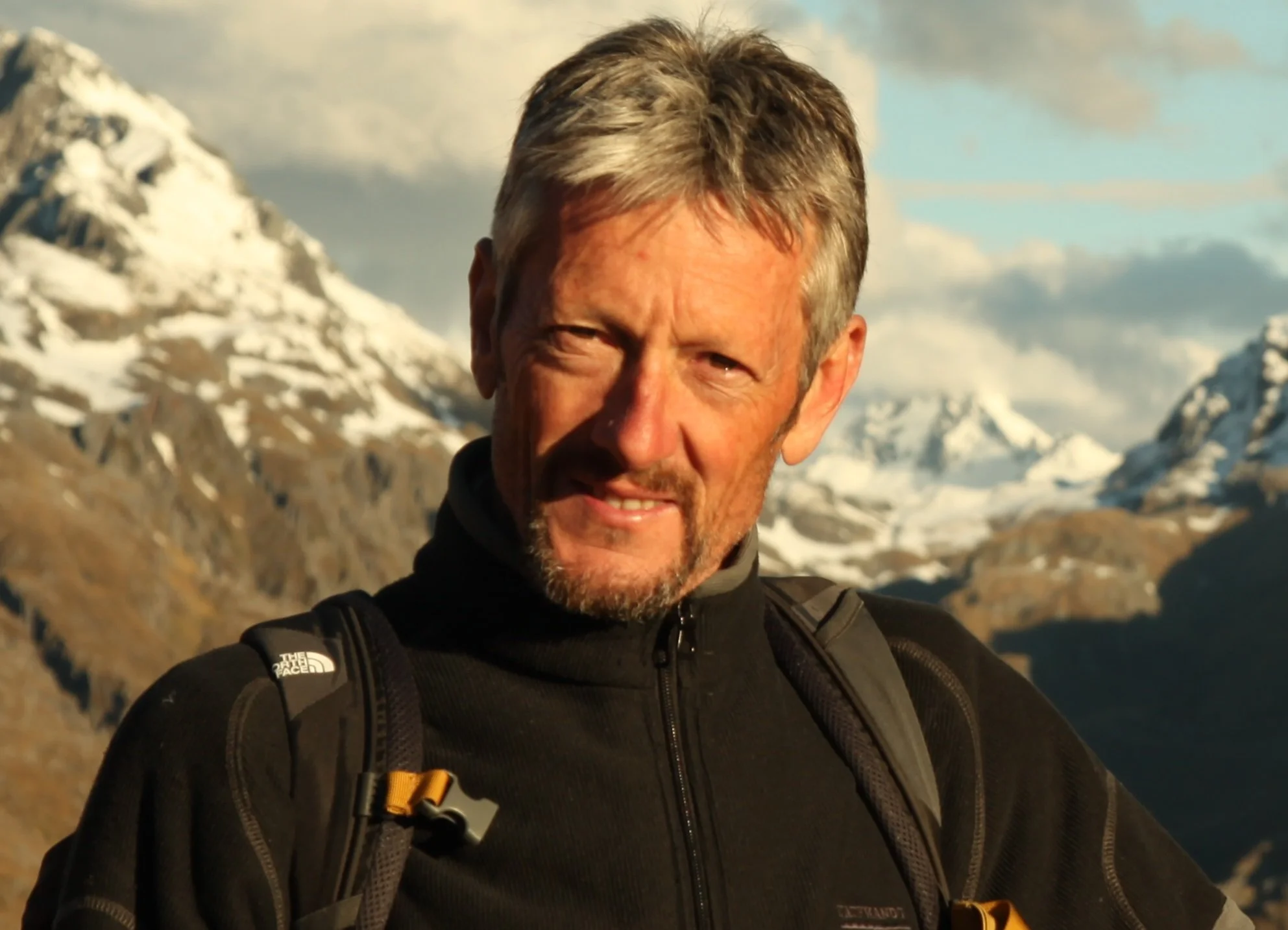 A middle-aged man with gray hair and a beard outdoors in a mountainous, snowy landscape during daytime, wearing a black jacket and a backpack.