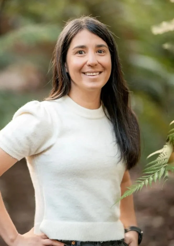 A person in a reflective vest operating a laser scanner or survey equipment under a concrete bridge, overlooking a landscape with trees and clear blue sky.