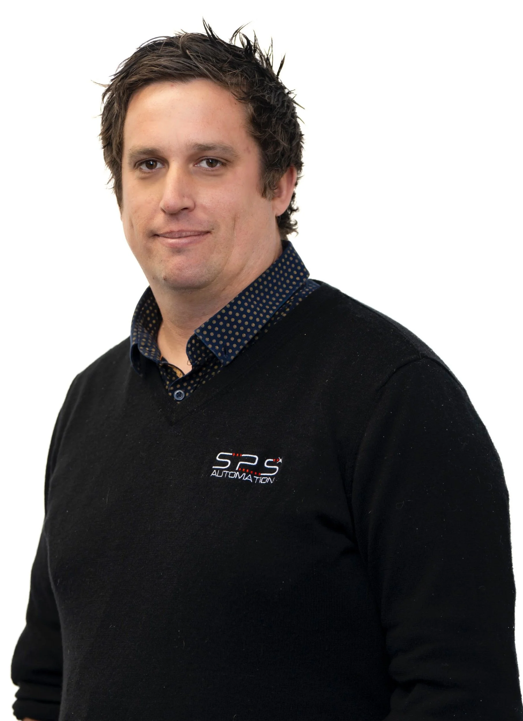 A man with dark brown hair and light skin, wearing a black sweater with a logo that says 'SPS Automation' over a dark blue, polka-dotted collared shirt, standing against a plain white background.