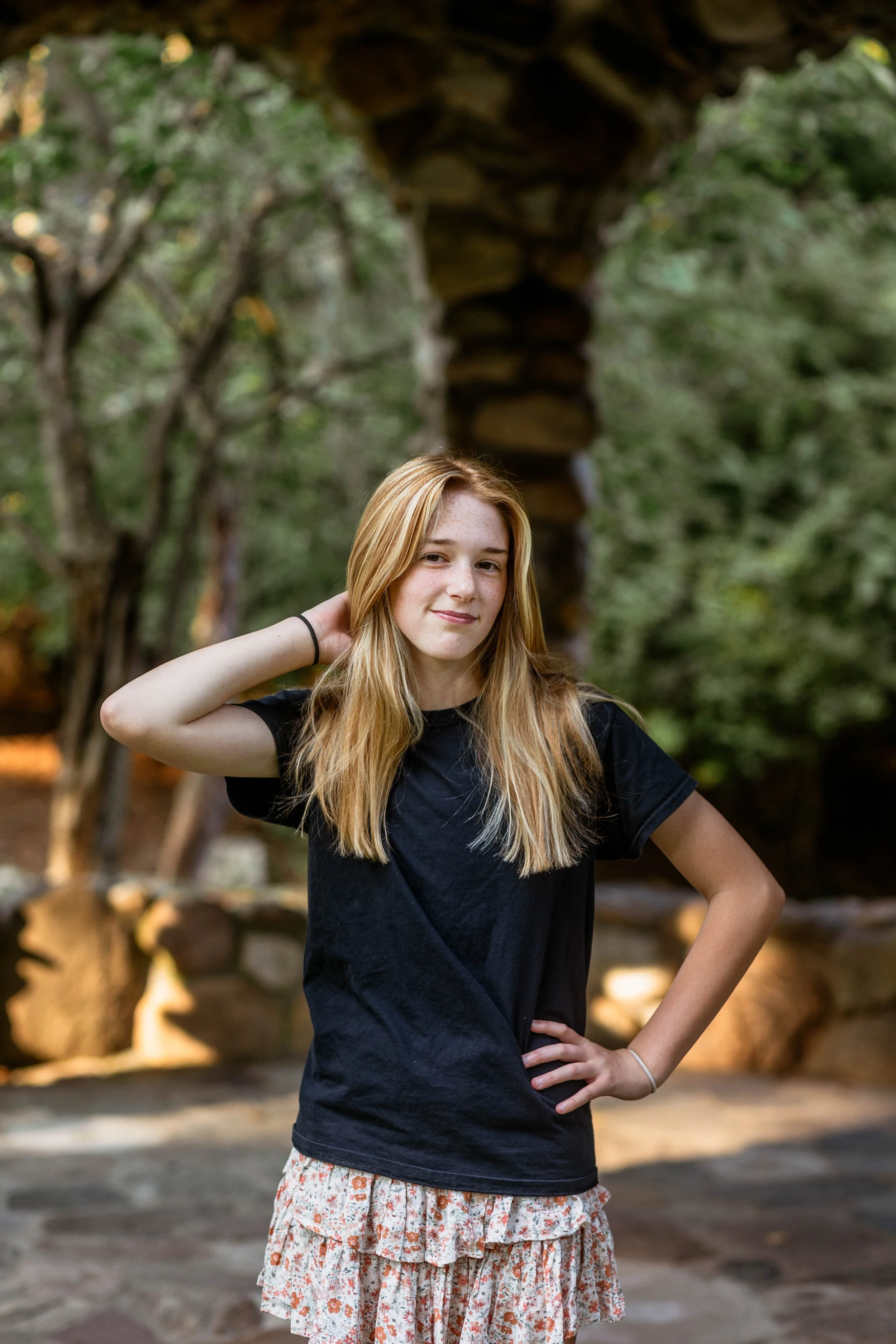 Young woman with long blonde hair wearing a black shirt and floral skirt, standing outdoors with a stone archway in the background.