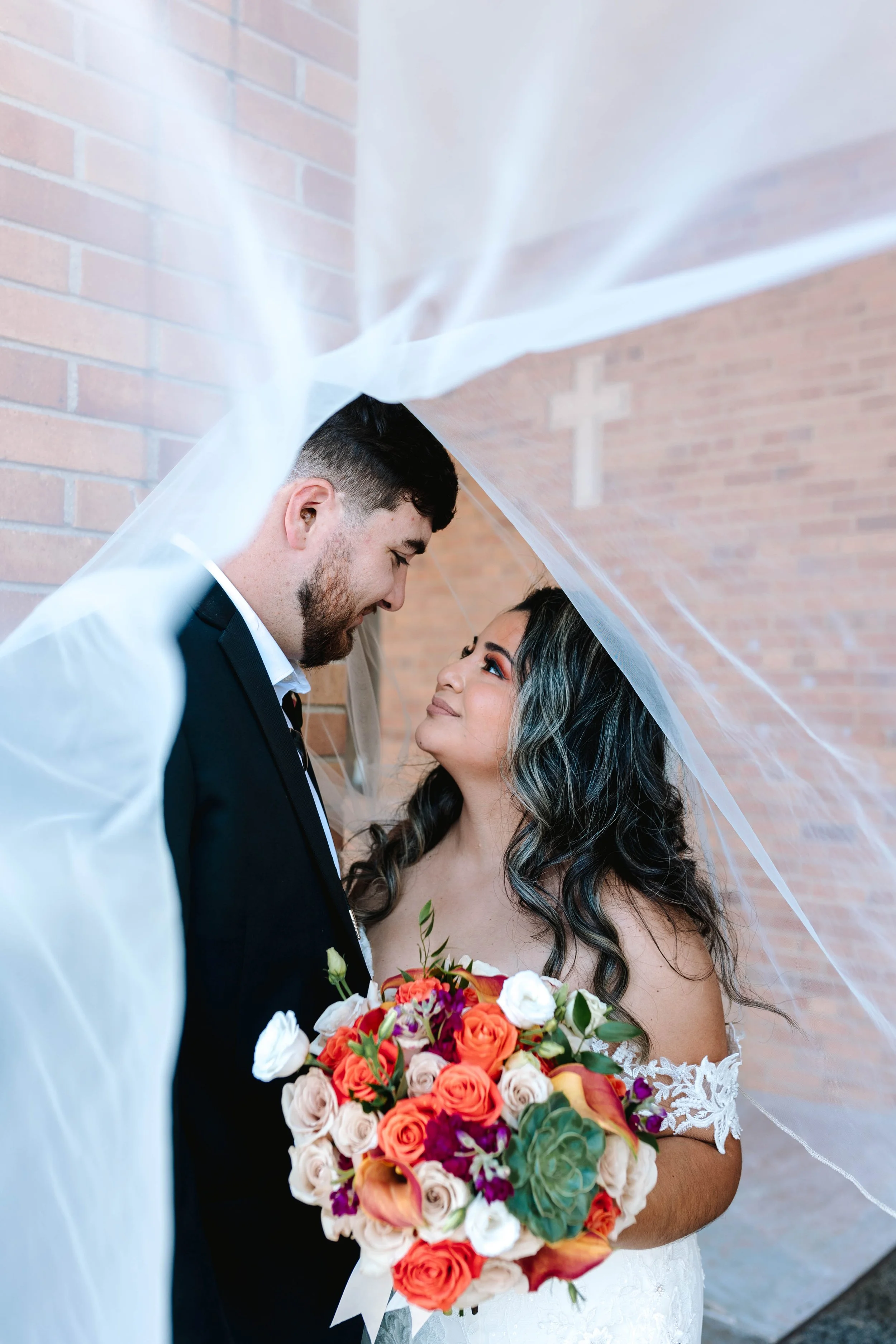 Bride and groom embracing under wedding veil, bride holding colorful bouquet, brick wall background, cross visible.